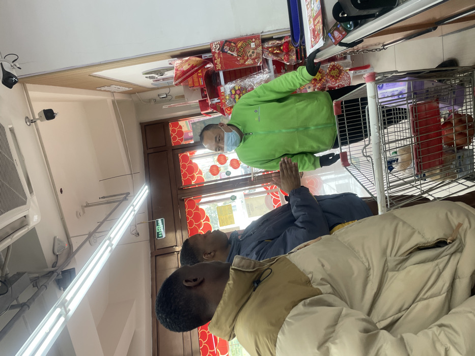 Inside a store decorated with red lanterns, two people wearing jackets are at the foreground, possibly customers. Behind them, a person in a green jacket wearing a face mask stands next to a shopping cart filled with items, possibly a store worker assisting customers. The store interior features a tiled ceiling, fluorescent lighting, and security cameras. Merchandise is visible on shelves behind the employee.