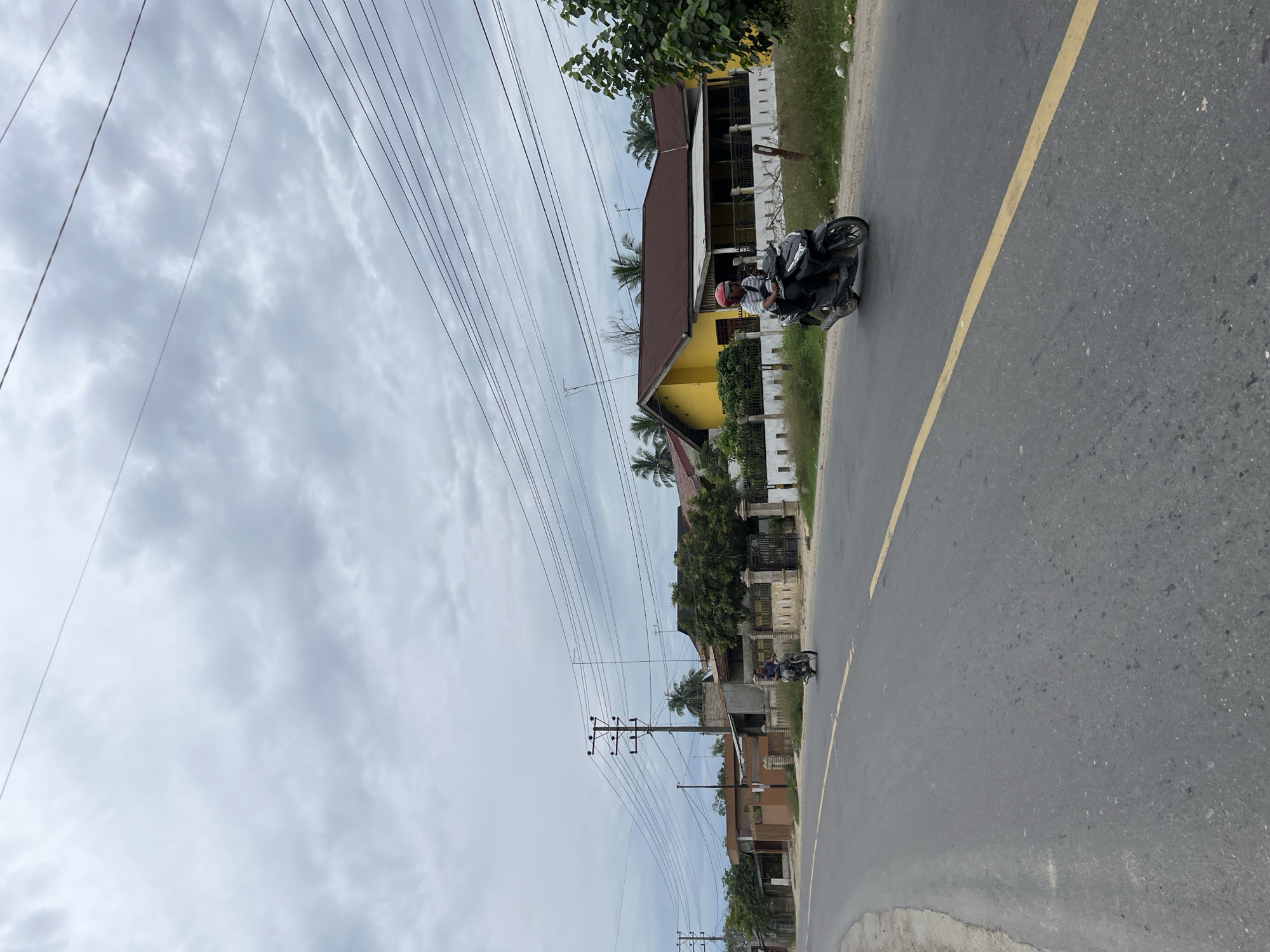 This is a vertical, eye-level, medium-range shot of a road in Simpangempat, Indonesia, with a cloudy sky overhead. The road is paved with asphalt and has a yellow dividing line. Two individuals on motorcycles are visible. In the foreground, on the right side of the road, a person wearing a red helmet is riding a black scooter, moving away from the camera. Further down the road, on the left, another person on a motorcycle is also moving away from the camera. The road is lined with houses and vegetation. Several utility poles with numerous electrical wires stretch across the upper portion of the image. The houses vary in color and style, with a prominent yellow house with a brown roof being a focal point on the right side of the road. Trees and fences are also present along the roadside. The overall impression is that of a typical residential street in a tropical climate.