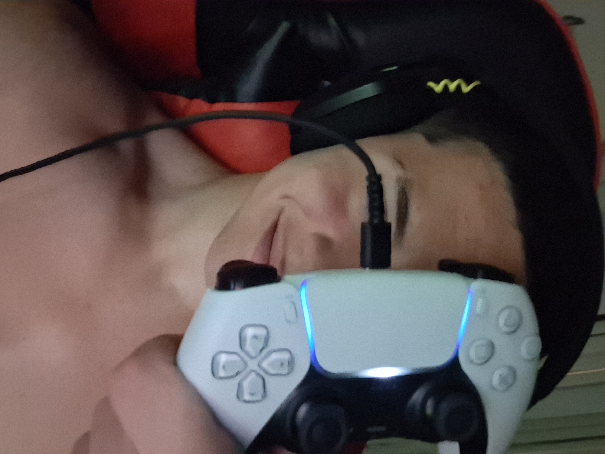 The image shows a close-up, vertically oriented, head-and-shoulders shot of a young person with short dark hair, wearing headphones, and holding a white video game controller in front of their face. The person is looking towards the camera with a slight smile. The controller is obscuring the right side of their face and eye. The background is dark and appears to be an indoor setting, possibly a gaming room, with a red chair visible. There is no information in the image that would allow for the determination of its location in Varna, Bulgaria.