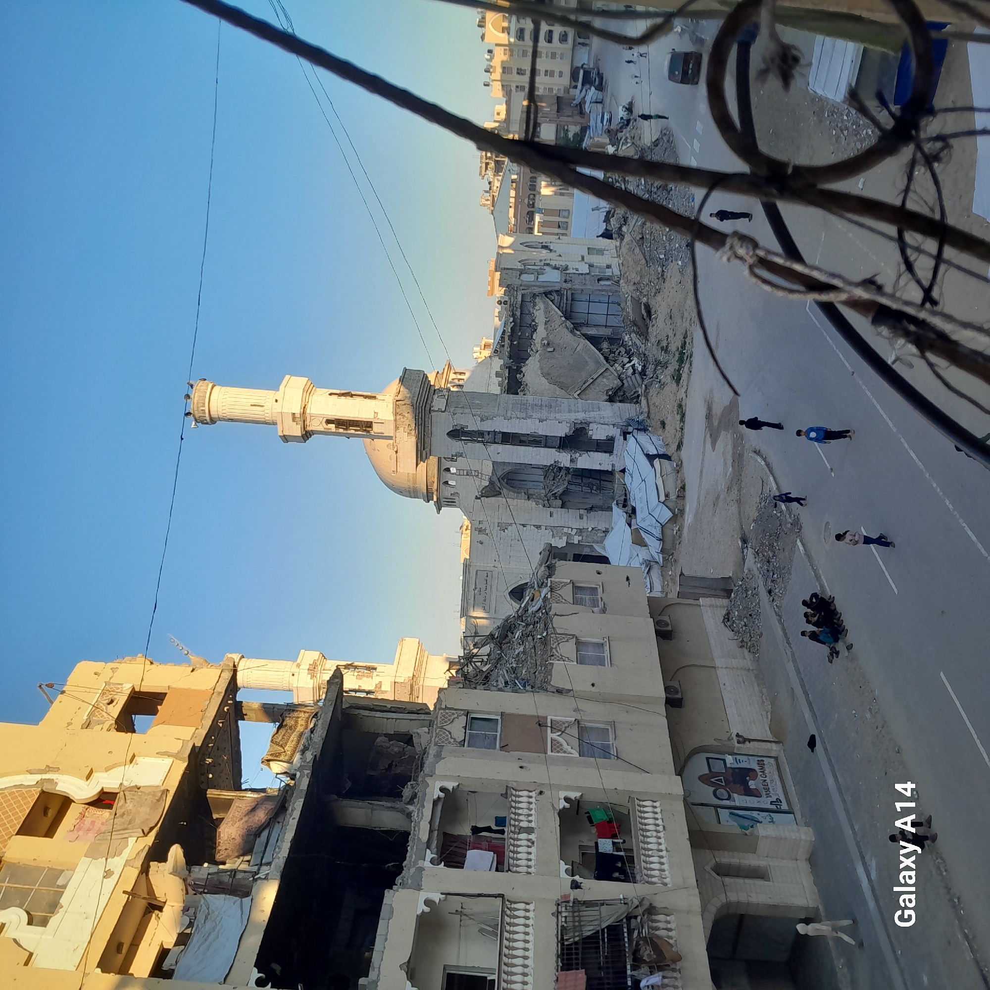 The image shows a scene of devastation in what appears to be an urban area. The location includes buildings that have sustained significant damage, with one building notably damaged, its floors and walls partially collapsed. A mosque with a minaret is also present, showing structural damage. A group of people are standing on the street beside the damaged buildings. Overhead, there are exposed wires. The photo seems to have been taken from an elevated position.