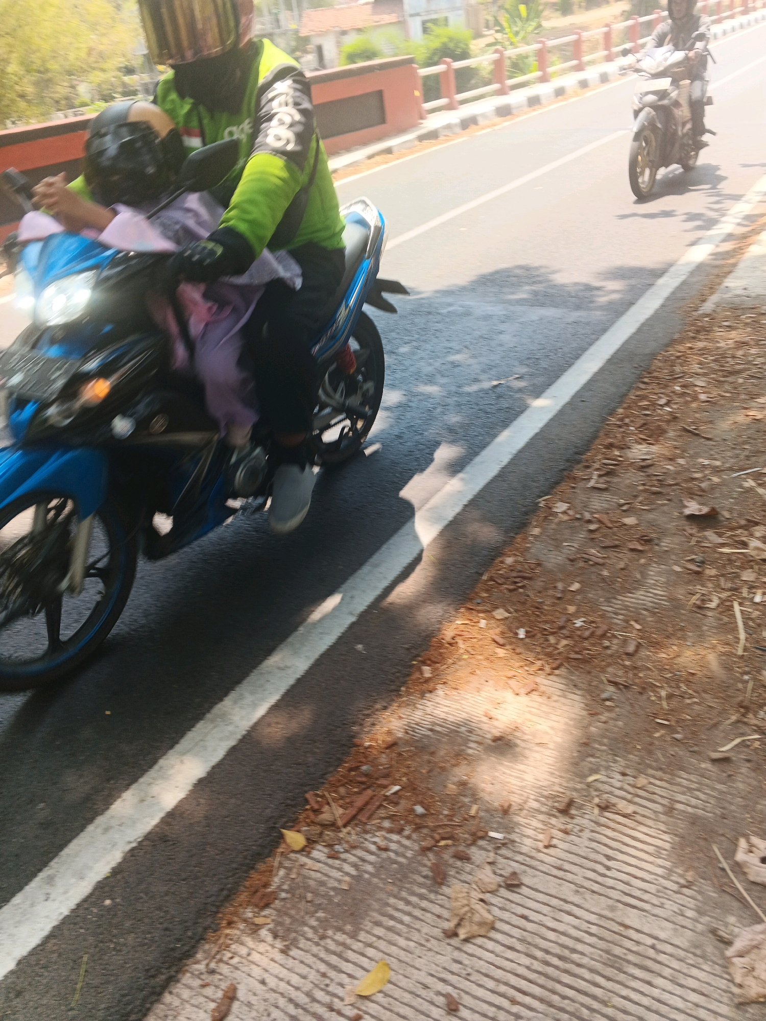 Two individuals are riding on a blue motorcycle, moving along a paved road. The person in the front is wearing a green long-sleeved shirt with black accents, a black helmet, and is holding onto the handlebars. The person behind them, appearing to be a child, is wearing a black helmet and a light purple garment. The motorcycle has its headlights on and is positioned on the left side of the road, with a white line marking the lane. The environment suggests an urban setting, possibly in Semarang, Indonesia, with a red and white barricade visible in the background, and trees lining the side of the road. Another motorcycle, ridden by a person in a black helmet, is visible further down the road. The road surface is asphalt, with loose debris and fallen leaves on the shoulder, which is constructed of concrete with visible parallel lines. The lighting indicates it is daytime, with shadows cast on the road.