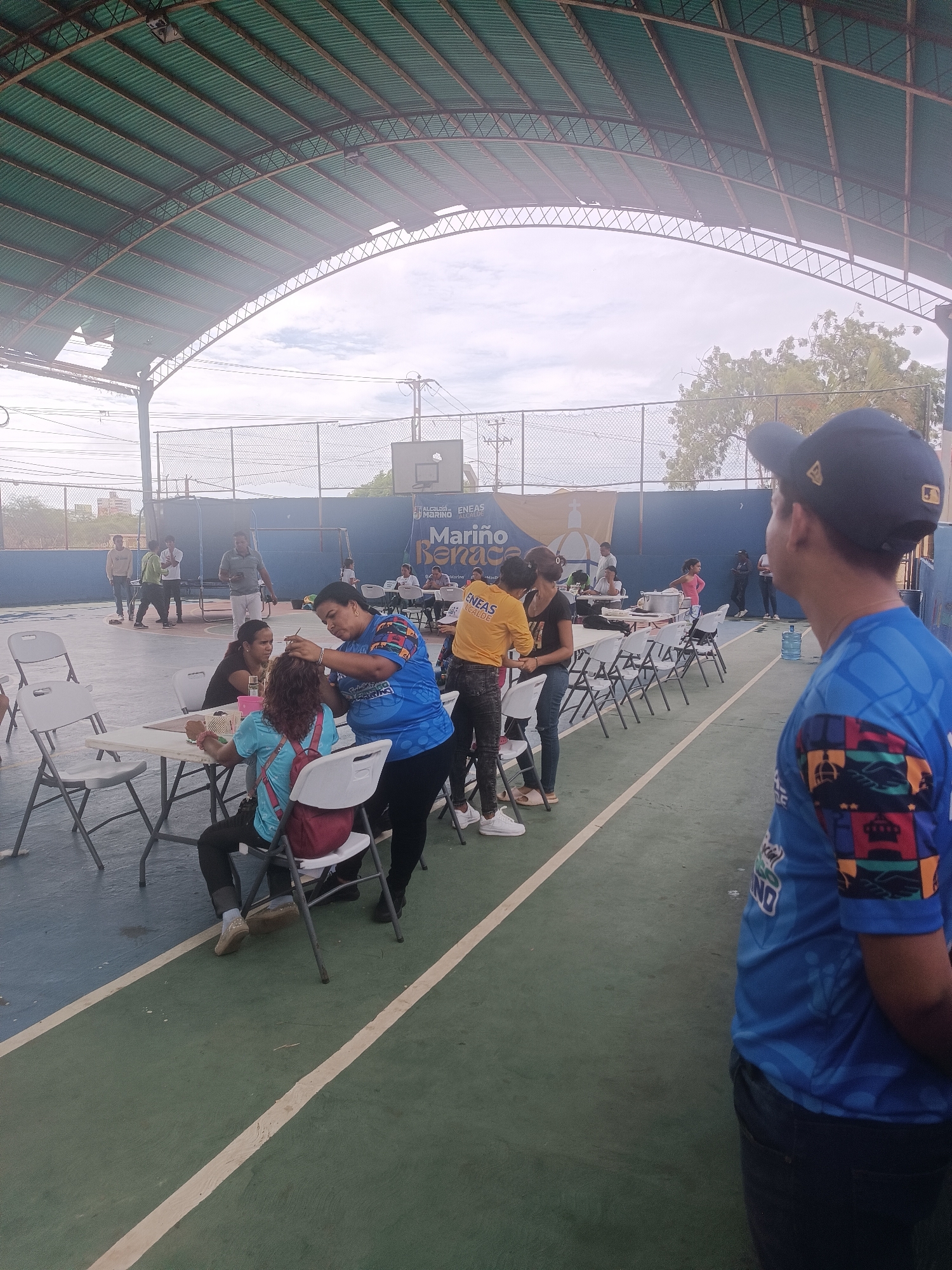 The image depicts an outdoor, covered sports court, likely a basketball court, in El Piache, Venezuela, with a community gathering in progress.
**Environment and Structure:**
The court features a green and blue painted floor with white dividing lines. An arched metal truss structure supports a green corrugated metal roof, parts of which show wear or damage. Light fixtures are attached to the roof structure. Beyond the court, a chain-link fence is visible, with dense green foliage and utility poles against an overcast sky. A blue wall, possibly part of the court perimeter, features a basketball backboard and hoop. A large blue and yellow banner is affixed to this wall, displaying text that includes "ALCALDÍA DE ENEAS MARIÑO ALCALDE" and an emblem resembling a domed building.
**People and Activities:**
Approximately fifteen to twenty individuals are present on the court. In the immediate foreground, facing away from the camera and towards the activity, stands a man wearing a dark blue baseball cap and a blue short-sleeved shirt with a multi-colored patterned design on the sleeves and dark trousers. To his left, a large transparent water dispenser bottle rests on a stand.
In the midground, several white plastic folding tables and chairs are arranged on the green section of the court. At one table, a woman in a blue t-shirt with a colorful pattern (possibly a uniform, with visible text "MARIÑO") is actively styling or braiding the hair of another woman seated opposite her. The seated woman has curly reddish-brown hair and is wearing a light blue top and a red backpack. Another woman is seated to the left of the woman receiving the hair treatment.
Further into the background, other individuals are standing and conversing or engaged in activities around additional tables. One woman is visible in a yellow t-shirt that reads "ENEAS ALCALDE." Several other individuals are standing near the fence line on the left side of the court, observing the scene or interacting among themselves. The overall scene suggests an organized community event or service being offered.