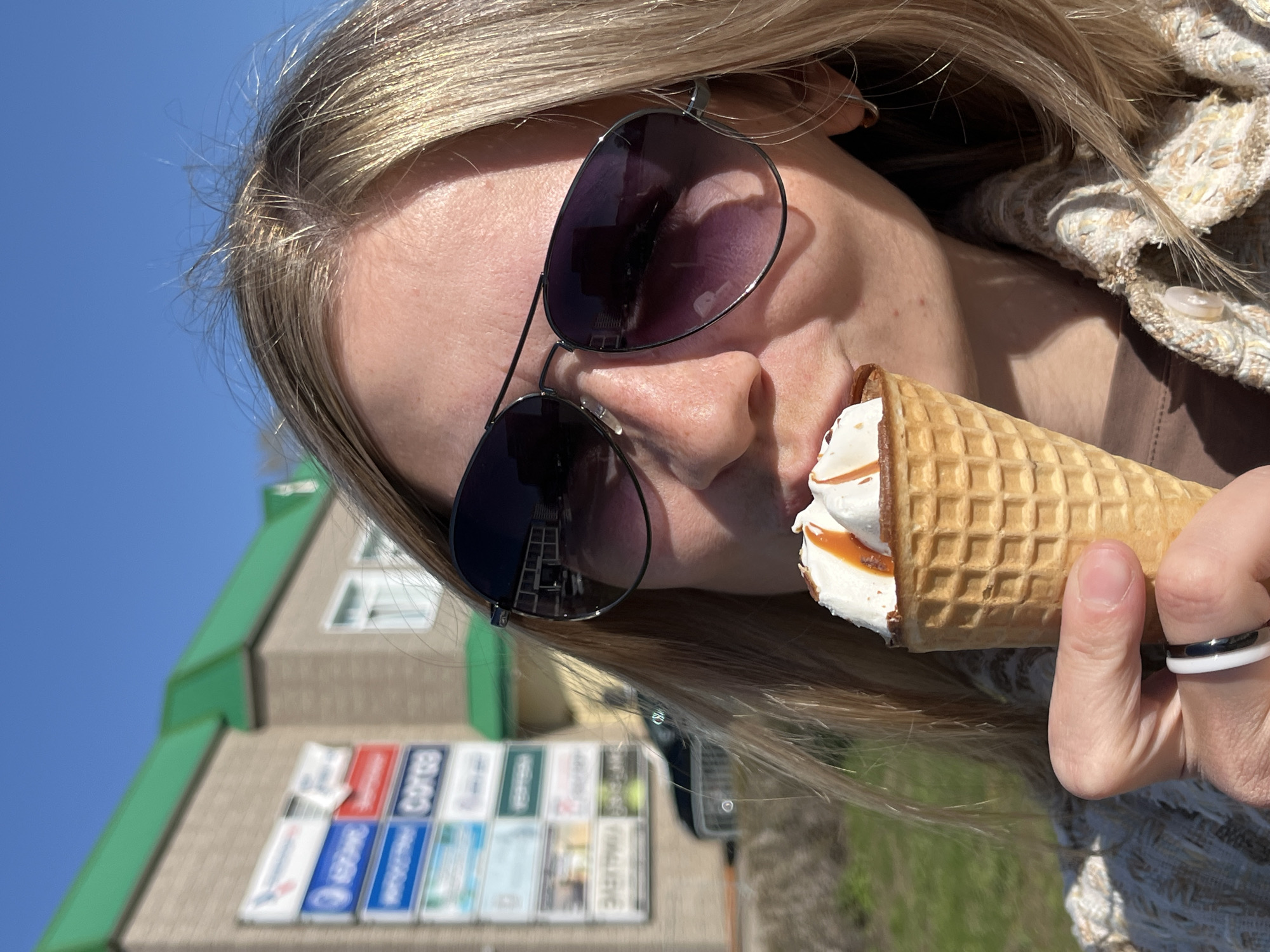A woman with long blonde hair, wearing sunglasses and a tweed jacket, is eating an ice cream cone. She is standing outdoors on a sunny day. In the background, there is a building with a green roof and a display of license plates. The license plates suggest this location is in the city of Chaikovsky, Russia.