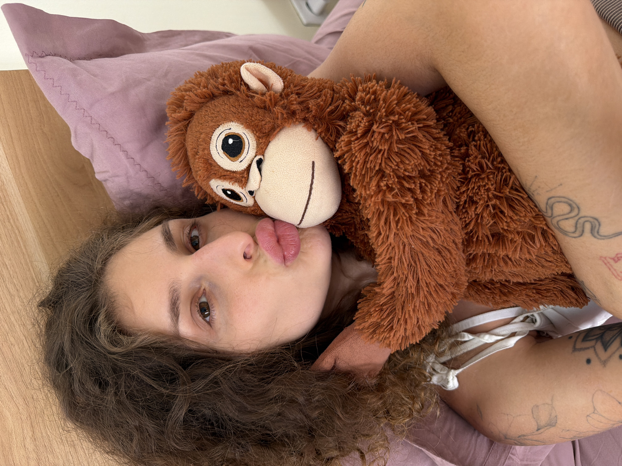 A person lies on their back on a bed with light purple sheets, holding a brown stuffed monkey. The person has curly brown hair, fair skin, and is puckering their lips. The monkey is positioned close to their face. The person's arms are visible and display tattoos. The location is presumed to be indoors, possibly a private residence. The location provided (North Kuta, Indonesia) cannot be verified from the image.