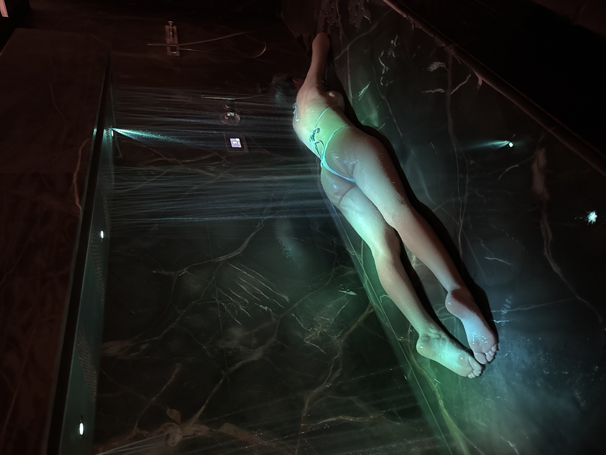 A female individual is depicted lying prone on a large, dark, polished marble slab within a dimly lit shower or spa room in the city of Unknown, Unknown. She is wearing a light-colored string bikini. Water streams downwards from an overhead shower fixture, with multiple jets forming sheets of water. A prominent green light illuminates sections of the falling water and casts a greenish glow across the individual's back, buttocks, and legs, which are visibly wet. The surrounding walls are also composed of dark, veined marble. Overhead, a rectangular panel contains multiple recessed white lights. Additional fixtures, including a metallic control panel and a small digital display, are mounted on the right-hand wall. The individual's head is turned away, and her arms are bent, resting on the surface.
