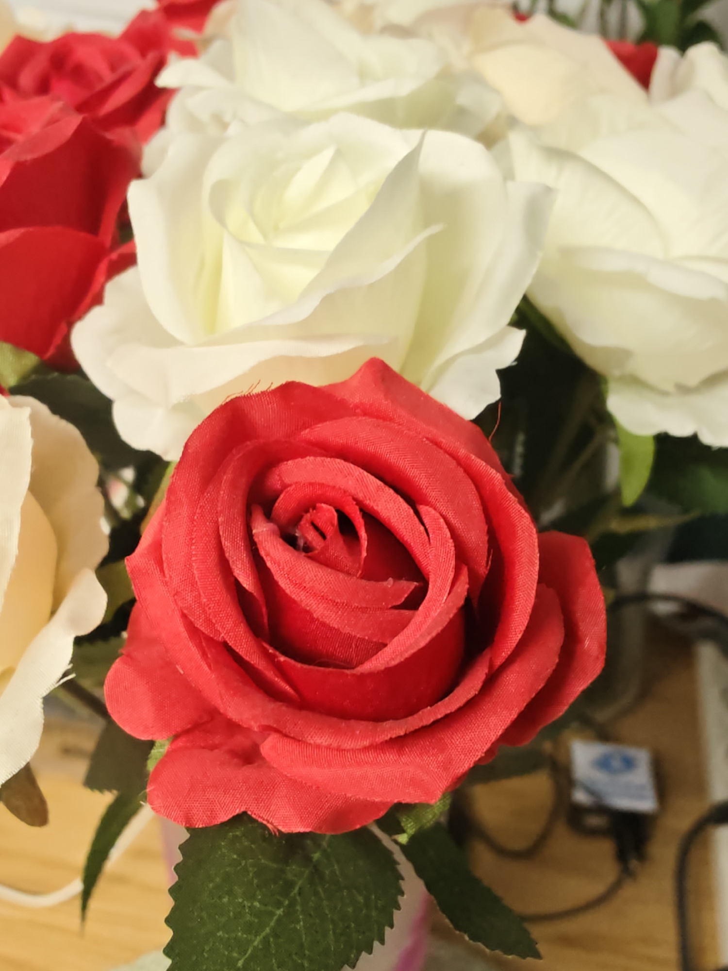 A close-up, eye-level shot features a bouquet of artificial roses in red and white, with one prominent red rose in the foreground. The bouquet is situated indoors, likely on a table, as indicated by the wooden surface visible at the bottom. In the background, partially blurred, are electronic devices and wires, suggesting a domestic or office setting. There is no indication of the specific location in Petah Tikva, Israel.