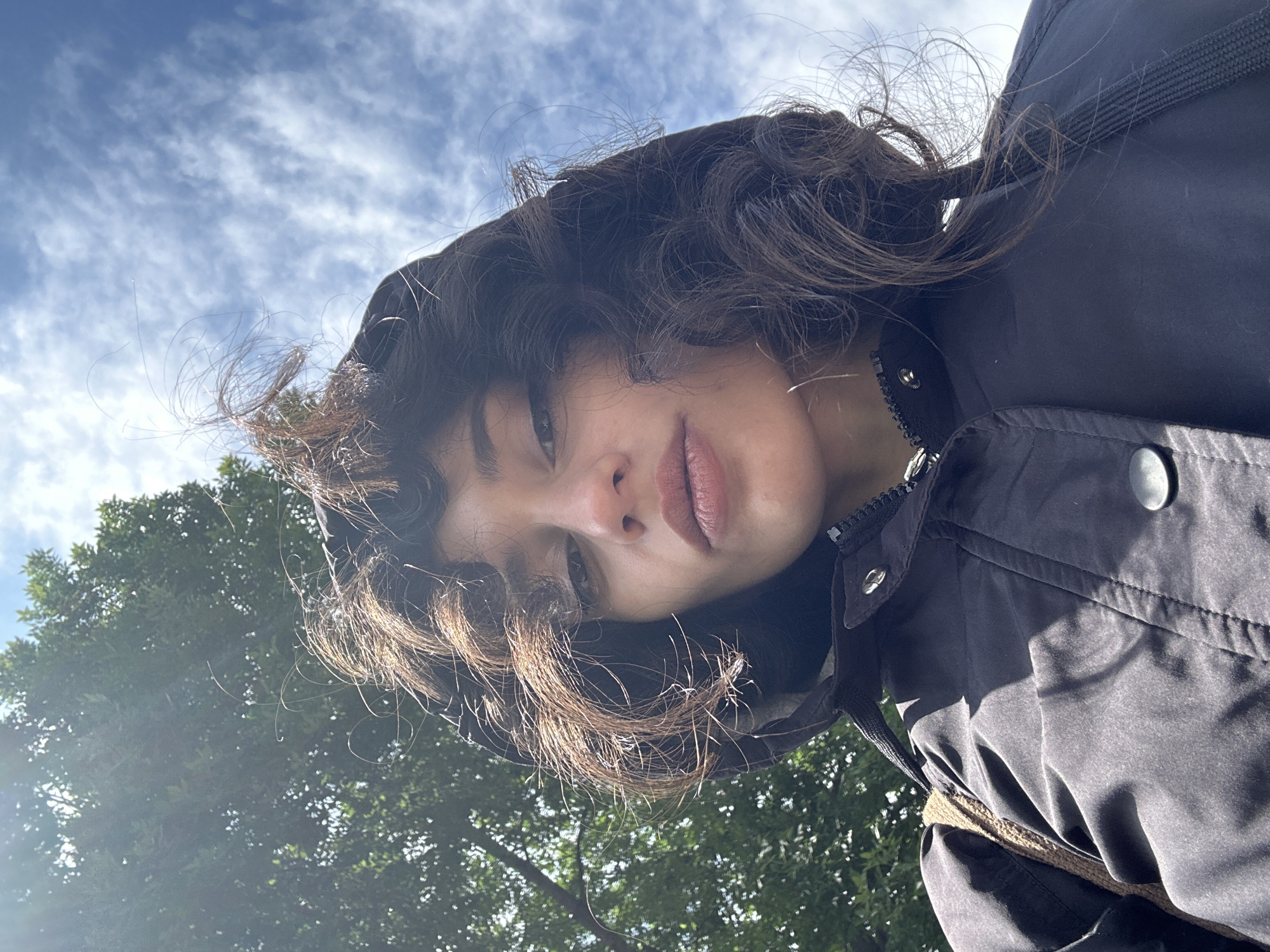 A young woman with dark, wavy hair is captured in a low-angle, upward shot, looking directly at the camera. She is wearing a dark hooded jacket, with the hood pulled up over her head. Her hair is windswept, with several strands blowing in the breeze. The background consists of a bright blue sky with scattered white clouds, and the green foliage of a large tree on the left side of the frame. The lighting suggests it is daytime, with sunlight filtering through the tree, creating some dappled light on the woman's jacket. The image appears to have been taken outdoors, likely in a park or a green area within the city of Sofia, Bulgaria, given the lush tree and the expansive sky.