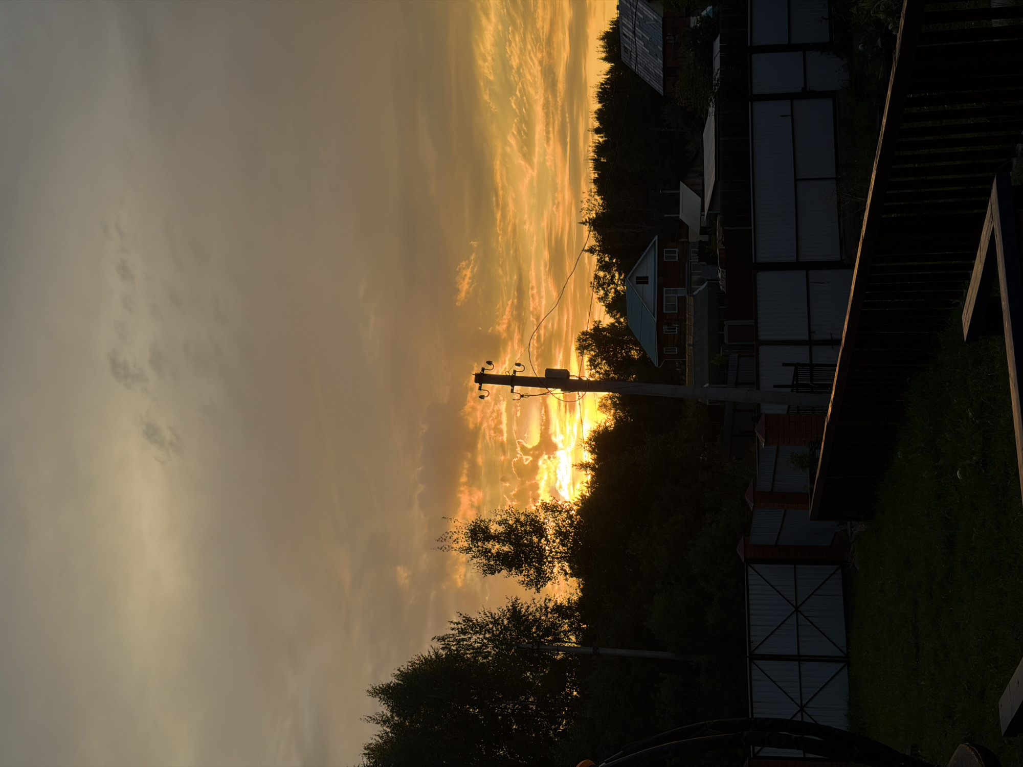 This image depicts a rural landscape at sunset, likely in the city of Chaikovsky, Russia, given the context provided. The sky is dominated by a dramatic sunset with vibrant hues of orange and yellow, illuminated by scattered clouds. In the foreground, there is a dark, grassy yard bordered by a wooden fence. A metal gate with a crisscross pattern is visible to the left, and a concrete fence with brick pillars extends to the right. Behind the fence, a line of trees forms a dark silhouette against the illuminated sky. Several residential buildings with dark roofs are nestled among the trees, and power lines are strung between utility poles. One prominent utility pole stands in the mid-ground, casting a shadow. The overall impression is of a peaceful, end-of-day scene in a suburban or village setting. There are no people or animals visible in the image.