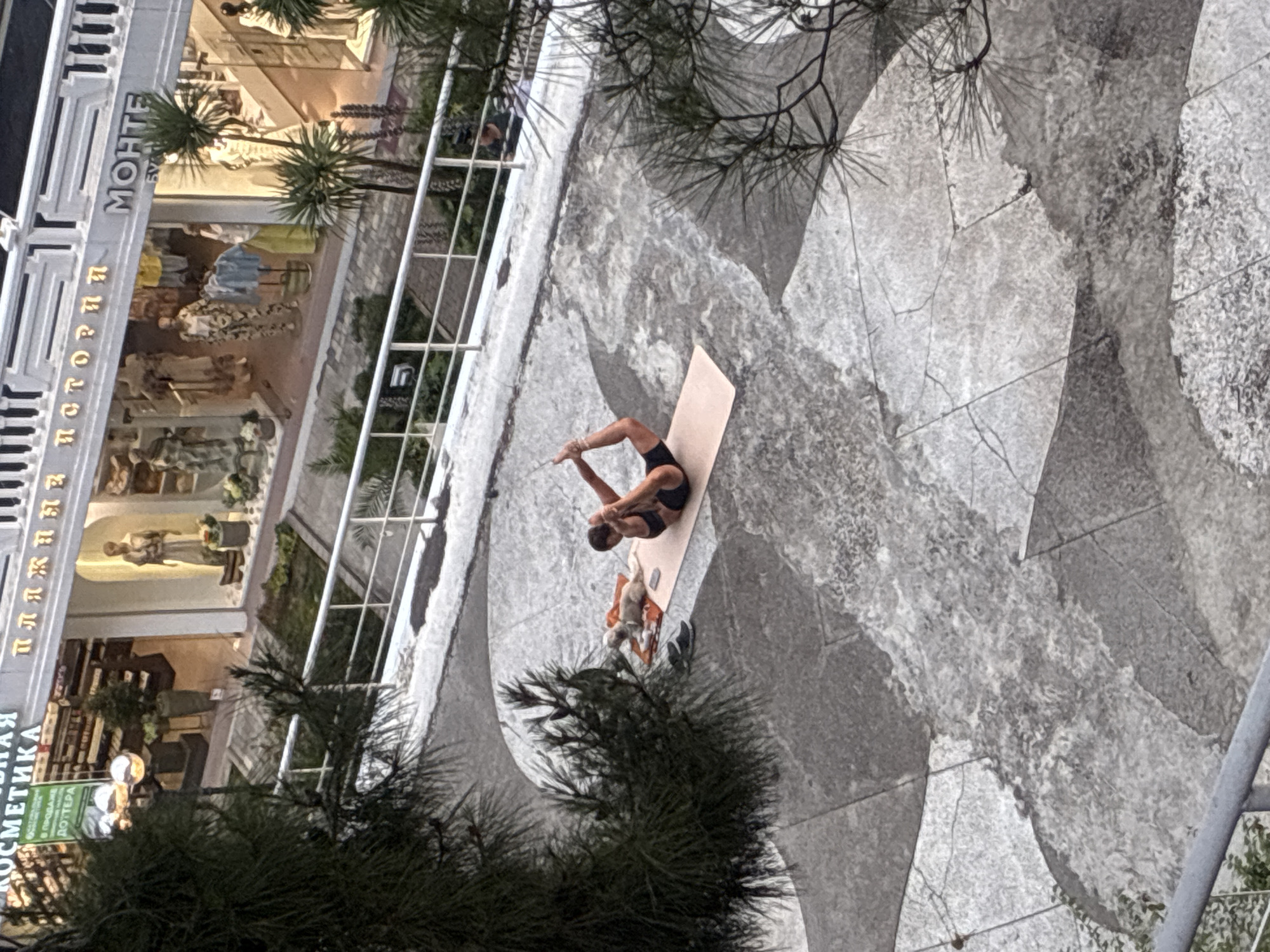 The image shows a person practicing yoga on a mat in an outdoor area. The person is in a backbend pose, with their legs and arms extended. A small dog is lying on a separate mat next to the yoga mat. The area appears to be paved with large stone tiles, with some cracked and worn sections. In the background, there is a building with storefronts, including one that appears to be a clothing store. The lettering on the storefronts is in Cyrillic. This scene is consistent with the coastal promenade in Sochi, Russia, which features shops and public spaces along the Black Sea.