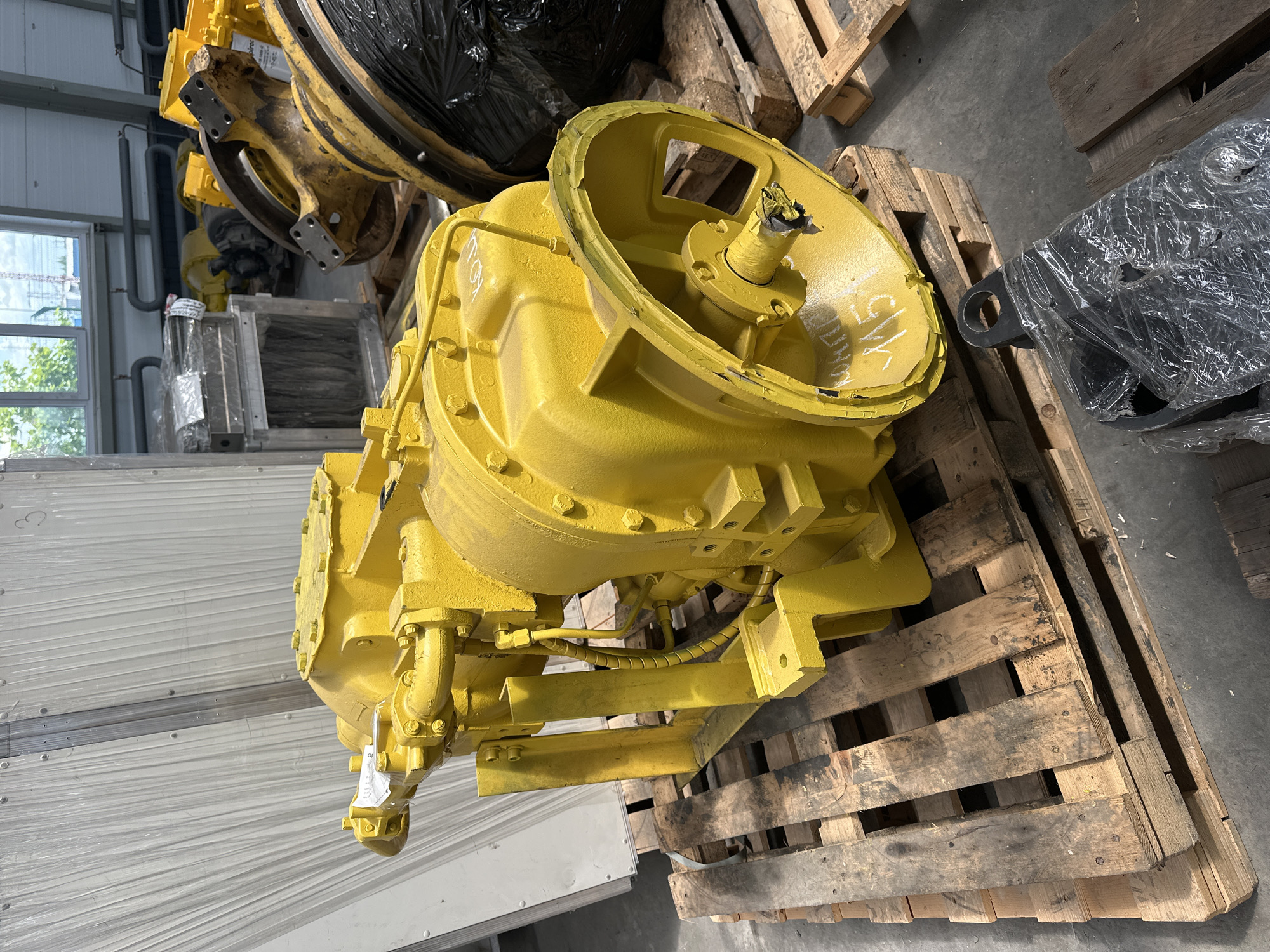 The image shows a bright yellow, heavy-duty mechanical component, possibly a gearbox or transmission, resting on wooden pallets. The object is highly detailed with pipes, bolts, and a large wheel-like structure on the right side. White markings, including "115", are visible on the yellow surface. The background reveals a warehouse or industrial setting with corrugated metal walls and other machinery, some wrapped in plastic. The scene is well-lit, suggesting an indoor environment. The presence of the mechanical component on pallets indicates it is likely stored or awaiting transport. The location is identified as Hora, Ukraine.