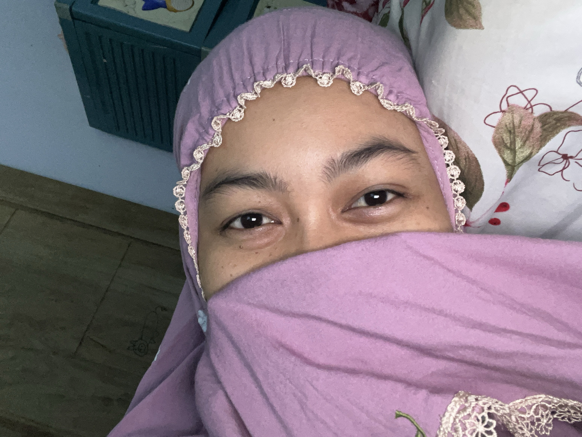 The image shows a close-up, head-and-shoulders shot of a young person lying down, partially covered by a lilac-colored garment, possibly a hijab or prayer garment. The person's face is turned towards the camera, with only their eyes and forehead visible. Their eyes are dark and open, and their eyebrows are well-defined. The garment has a delicate lace trim along the edge that is visible near the face. The background includes a textured wall and a green cabinet with horizontal slats. The person is lying on a pillow with a floral pattern. The lighting suggests an indoor setting, possibly a bedroom. The scene appears to be a candid or informal photograph. There is no specific information within the image to pinpoint its location in Klapanunggal, Indonesia.