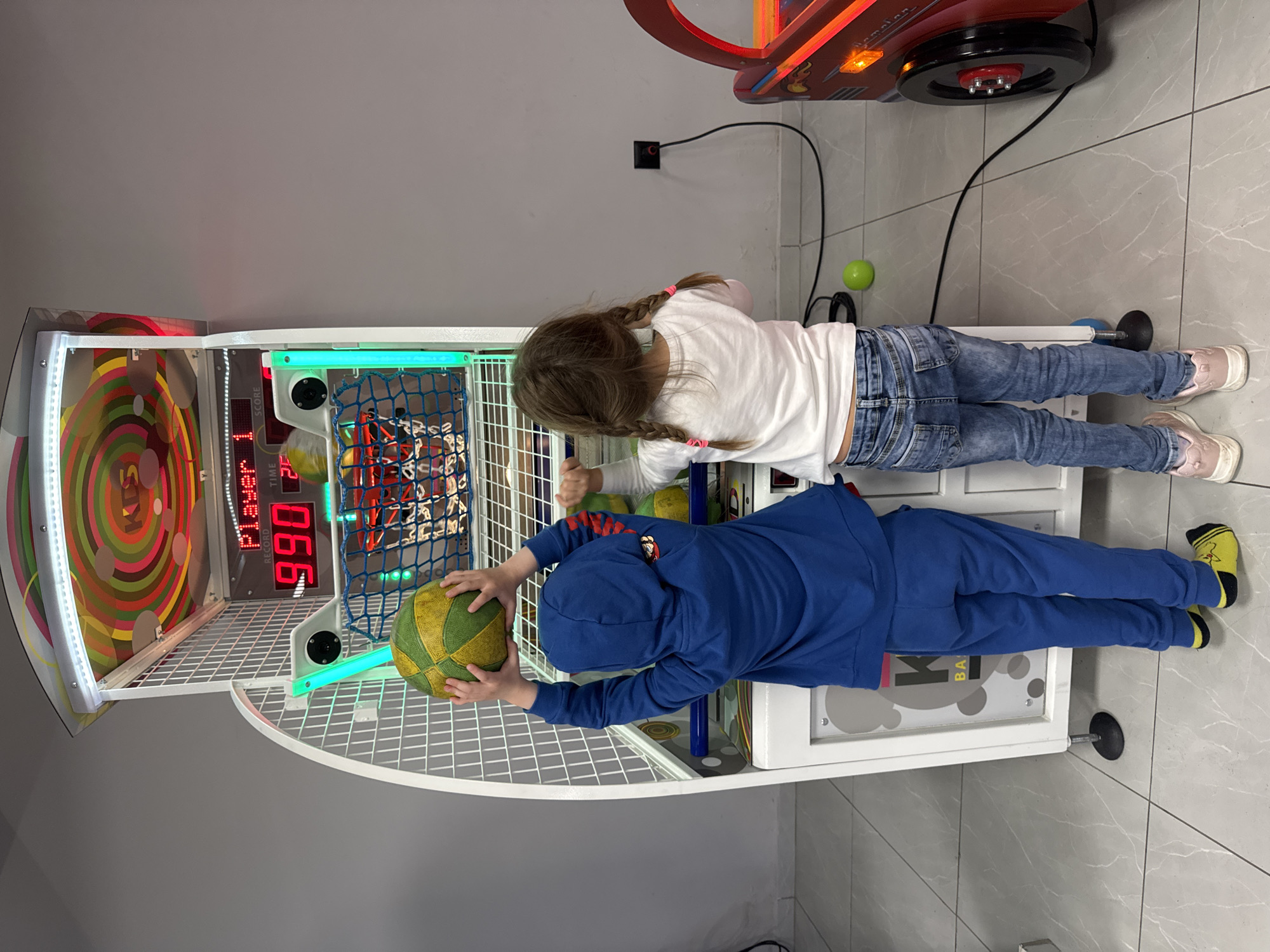 Two children are playing a basketball arcade game indoors. A girl with pigtails and wearing a white shirt and blue jeans stands behind a boy wearing a blue hooded sweatshirt and blue pants. Both are holding a green and yellow basketball, presumably to throw it into the hoop. The arcade game has a digital display showing the score. The surrounding environment has tiled floors and gray walls. Next to the game is a ride with lights and a steering wheel. The precise location in Kranevo, Bulgaria, is not identifiable from this image.