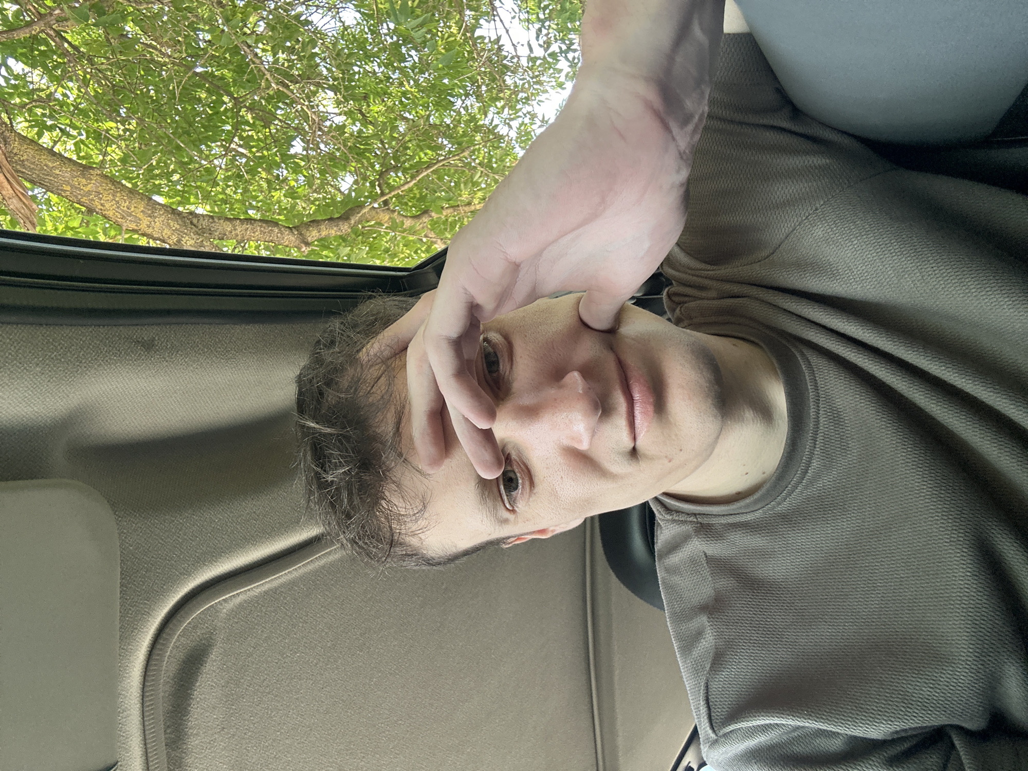 A selfie taken from inside a vehicle shows a Caucasian male in his late 20s or early 30s. He has short, brown hair and light skin. His right hand is raised to his forehead, obscuring his eyes. He is wearing a grey, textured long-sleeved shirt. The interior of the car is visible, with a grey headliner and a dark headrest. Outside the vehicle, through the window, a tree with green leaves is visible against a bright sky. The image appears to have been taken during the daytime, likely in a rural or suburban setting, as suggested by the presence of the tree. The specific location in Makiv, Ukraine is not discernible from the visual information presented in the image.