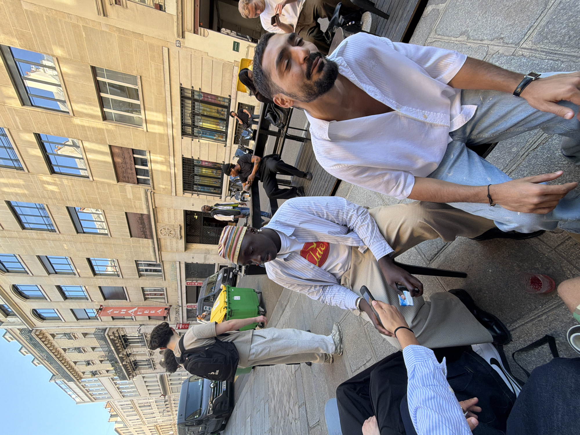 The image captures an outdoor scene in Paris, France. Several people are seated at tables, engaging in conversations and using mobile phones. A man with a beard and a partially unbuttoned white shirt is in the foreground. Another man wearing a striped hat and a white t-shirt with a logo is looking at a mobile phone. The surrounding environment includes buildings with windows, a green trash bin, and parked cars along the street. A person with a backpack walks past, near the bins. A red banner hangs between the buildings.