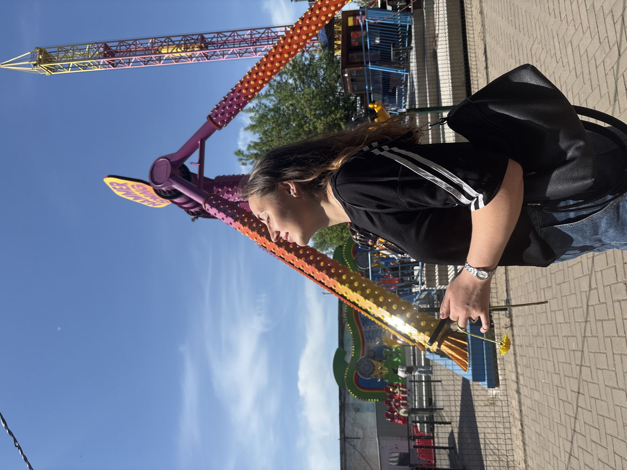 A sideways view of a person holding a dandelion in front of an amusement park ride. The person is wearing a black t-shirt with white stripes, blue jeans, and a wristwatch. A black bag is slung over their shoulder. The amusement park ride, identified as "Hammer" in Russian, has a purple and orange arm. Other rides and trees are visible in the background. The ground is paved with light-colored bricks. The sky is blue with some white clouds. This image was taken in Izhevsk, Russia.