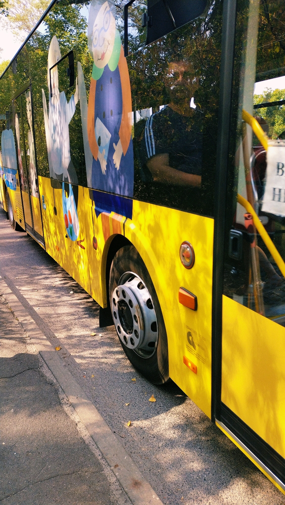 A yellow bus with cartoon graphics is parked on a street in Horishni Plavni, Ukraine. The bus has large windows that reflect the surrounding trees and sky. The graphics on the side of the bus depict cartoon characters. The bus is yellow with black trim and a large wheel is visible. The street is paved and a concrete curb runs alongside it. Fallen leaves are scattered on the ground. The lighting suggests it is daytime.