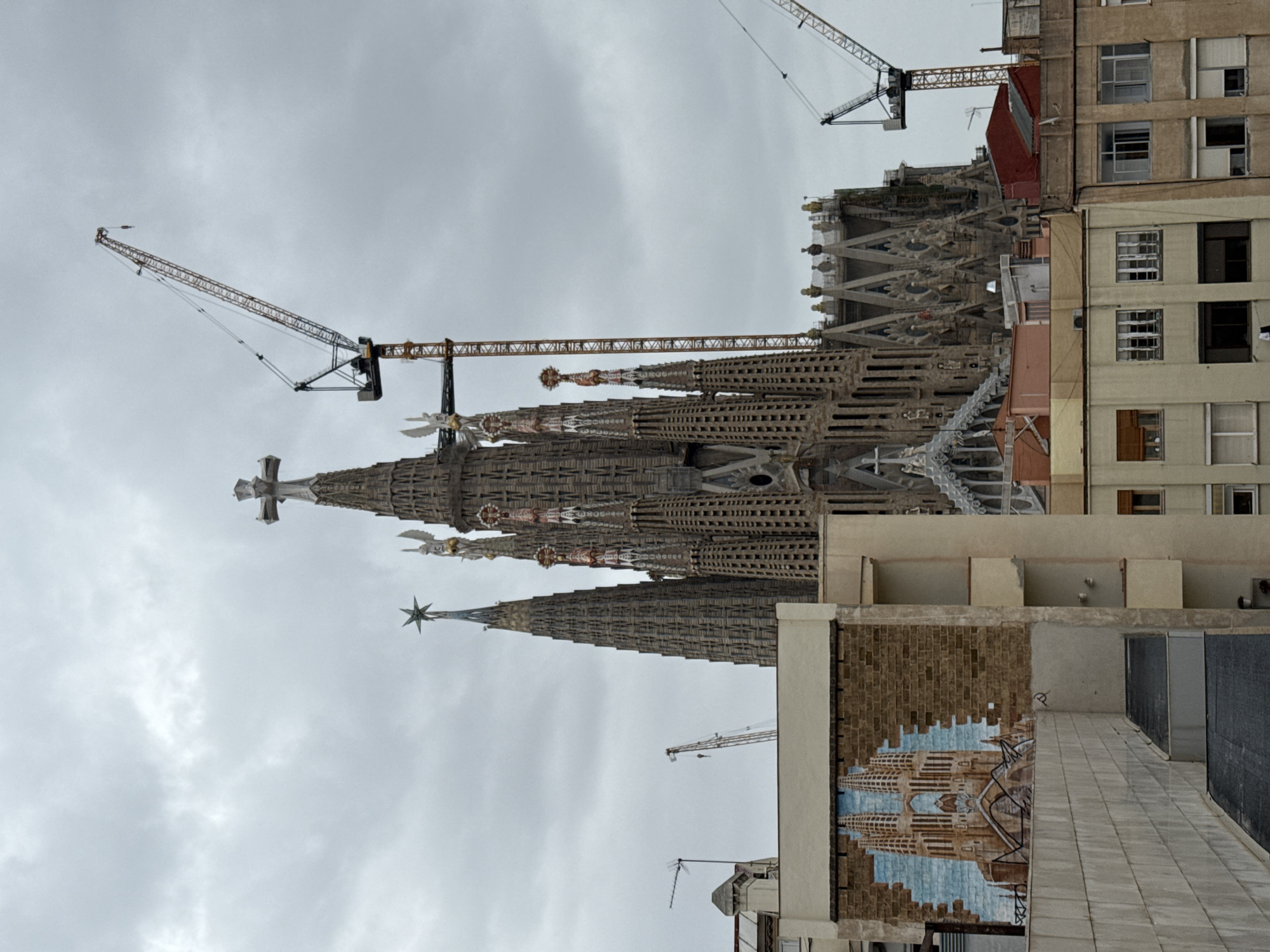 The image shows the Sagrada Familia in Barcelona, Spain. The iconic spires are under construction, as evidenced by multiple cranes. The sky is overcast. Adjacent buildings are visible, including an apartment building with several windows. A mural depicting the Sagrada Familia is on the side of another building.