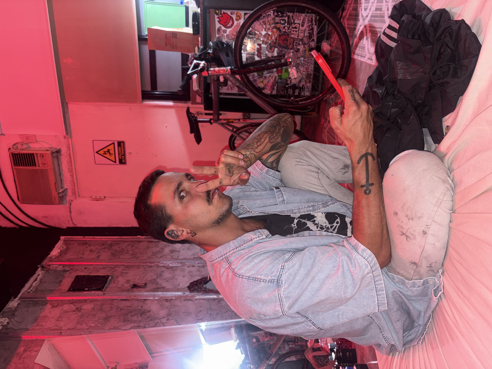 The image features a young man lounging indoors, illuminated by a dominant red light, suggesting an evening or nighttime setting. He is positioned on his side, facing the camera with a direct gaze and a neutral to slightly playful expression. His left hand is raised, making a peace sign, while his right hand holds a red pen or stylus. He wears a light blue denim shirt over a black graphic t-shirt and light-colored pants. Visible tattoos include a detailed sleeve on his left arm and an anchor-like symbol on his right wrist. Multiple earrings are also visible on his ears.
The background shows an eclectic interior. Behind the man, a bicycle stands, its wheel densely covered with various stickers. Next to the bicycle, a black jacket is casually placed on a soft surface, likely a bed or couch. To the left, an air conditioning unit is mounted on the wall. Below it, a yellow triangular warning sign is prominently displayed, featuring a black lightning bolt symbol and the Spanish text "PELIGRO ALTA TENSION" (Danger High Voltage), which is a location cue for Argentina. The overall scene depicts a casual and relaxed moment in a personalized urban space.