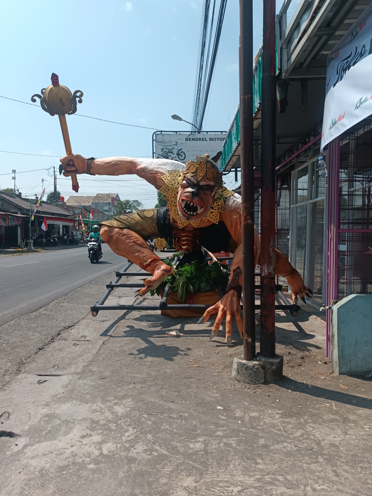 An oversized, animatronic ogre-like figure with an orange complexion, sharp teeth, and a golden headdress is displayed on the sidewalk next to a road in Imogiri, Indonesia. The creature is positioned as if lunging forward, with its left arm raised, holding a golden mace-like object, and its right arm extended with sharp claws reaching out. It appears to be part of a roadside display, possibly for a festival or local event, with banners and other businesses visible in the background. A person on a motorcycle drives by on the road, and the scene is bathed in bright sunlight, casting shadows on the pavement. The environment suggests a typical urban or semi-urban setting with buildings, power lines, and a paved road.