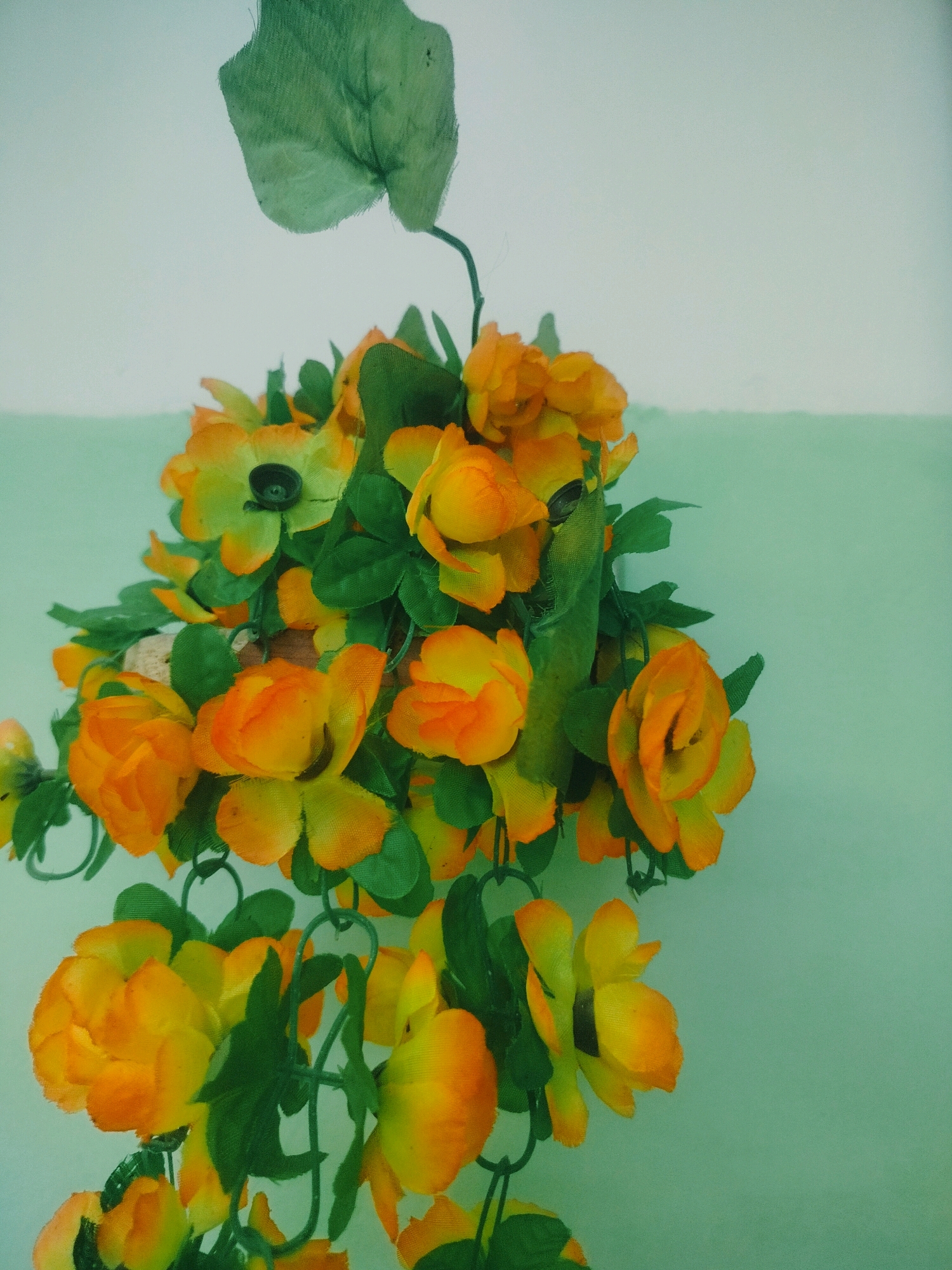 The image shows a close-up of a decorative garland of artificial orange and yellow flowers with green leaves. The garland is hanging against a two-toned background; the upper portion is white, and the lower portion is a light green. The flowers appear to be made of fabric. There are no people or animals visible in the image. The lighting is diffused. The scene appears to be indoors, possibly in a room or on a balcony. The provided location of Көлкент, Kazakhstan, is not directly ascertainable from the visual information presented in the image.