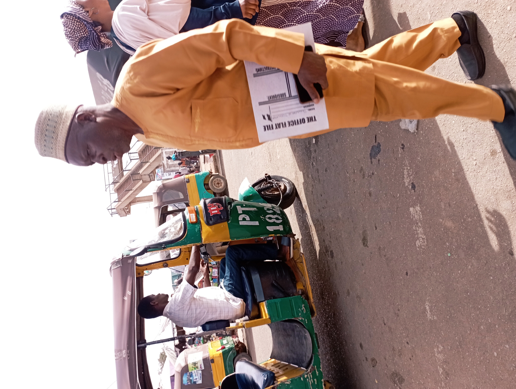This photograph, likely taken in Jos, Nigeria, captures a bustling street scene in bright daylight. The primary subject is a man in the foreground, seen from the side, walking or standing on a paved road. He wears a mustard yellow long-sleeved suit and trousers, with black loafers, and a light-colored, patterned kufi cap on his head. He holds a stack of papers and a black smartphone in his left hand. The papers display text, notably "THE OFFICE FLAT FILE" and "SUBSEQUENT INSTRUCTIONS," along with fields for "ADDRESS:" and other illegible details.
Behind the man, partially visible, is a woman wearing a patterned headscarf and clothing. Further back, a green and yellow three-wheeled auto-rickshaw, commonly known as a keke napep in Nigeria, is visible. It has several passengers inside, including a man in a white checkered shirt seated behind the driver, and another person seen from the side. On the rear of the tricycle, "PT 1899" is clearly inscribed in green capital letters.
The setting appears to be a sunny day, indicated by the strong shadows cast on the asphalt road. In the far background, elevated concrete structures suggest an overhead bridge or building. The overall scene depicts a typical urban environment with daily commute or pedestrian activity in Nigeria.