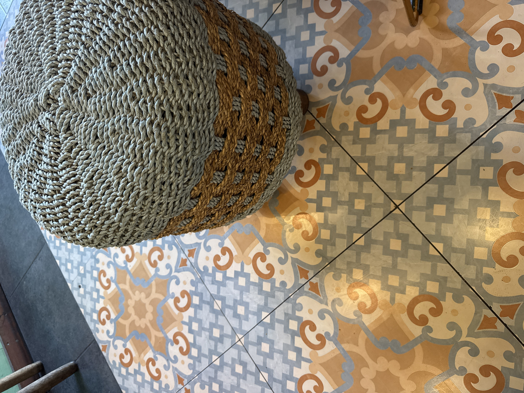 The image captures an overhead view of a woven pouf on a patterned tiled floor within an interior setting.
The main subject is a large, round pouf, likely made from natural fibers such as seagrass or jute. Its top surface features a tight, concentric spiral weave in a light greyish-beige color. The visible side of the pouf displays a contrasting design, incorporating both the greyish-beige and a darker brown fiber, woven into a patterned band that runs vertically around its circumference.
The pouf rests on an intricately patterned floor composed of square tiles. These tiles feature a repeating geometric and floral design in a palette of light blue, orange-brown, cream, and beige. The pattern includes swirling scroll motifs, small square grid-like sections, and larger, star-like formations where four tiles meet. Black grout lines separate the individual tiles.
In the lower-left corner, a section of darker, plain flooring is visible alongside what appears to be the wooden legs of another piece of furniture or a ladder, partially out of frame. The lighting suggests an indoor daytime scene, with soft, diffused light illuminating the space. No activity is taking place, and no text is visible in the image. The aesthetic, characterized by natural textures and intricate tile work, is consistent with interior design styles found in locations like Kuta Selatan, Indonesia.