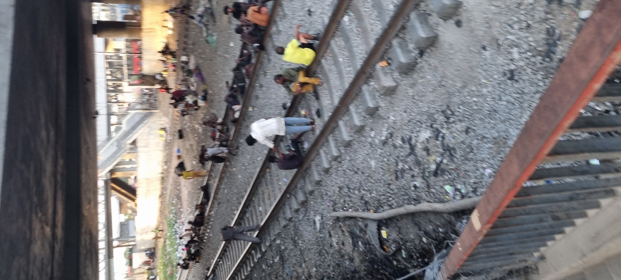 The image displays a detailed view of a scene located under an elevated overpass in an unidentified urban area. Multiple sets of railway tracks with concrete sleepers traverse the ground, which is composed of gravel and is heavily strewn with litter and debris.
**People:** Approximately 20 to 30 individuals are observed throughout the scene. Many are seated or lying directly on the railway tracks or on the ground immediately adjacent to them. In the mid-ground, a person wearing a white shirt and blue pants stands over another individual, dressed in a dark shirt, who is seated on the tracks; this interaction appears focused. To their right, two other individuals sit on the tracks. Further left on the tracks, a person in dark clothing walks. In the background, numerous other individuals are scattered, mostly seated. Their posture suggests resting or gathering.
**Objects:** A rusty metal railing with vertical slats occupies the lower foreground, indicating the viewpoint is from an elevated position. The railway infrastructure includes steel rails and concrete sleepers. The ground is covered in gravel, intermingled with discarded items such as plastic bags, fabric scraps, and various refuse. A long wooden log lies parallel to the leftmost track. In the background, an elevated pedestrian bridge with a concrete staircase is visible, alongside concrete support structures and metal fencing. Distant, rudimentary buildings are also discernible.
**Environment:** The setting is an urban transportation corridor beneath an overpass. The area exhibits significant neglect, characterized by widespread litter. The natural light source, possibly late afternoon or early morning sun, illuminates parts of the scene while the overhead structure casts shadows. The arrangement of individuals and their belongings indicates a temporary gathering or habitation within this industrial landscape.
