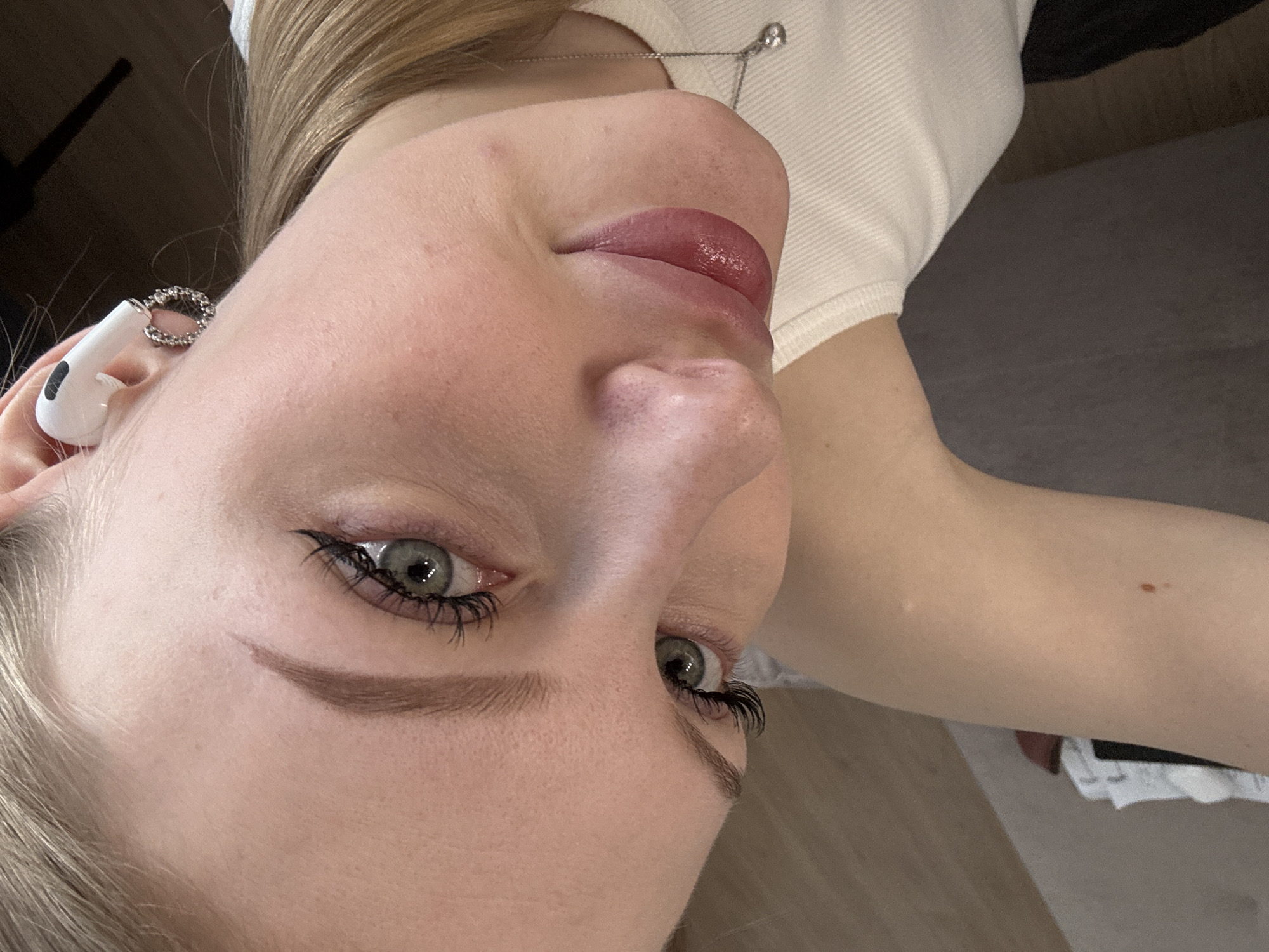 A close-up portrait of a light-skinned person with blonde hair. The person is wearing a white ribbed top and a silver necklace with a small pendant. An earbud is visible in the left ear, adorned with a sparkling ring. The person's face is angled towards the camera, showing prominent features: pink lips, a nose, and eyes with defined eyebrows and eyelashes. The background is indistinct, with hints of a dark surface. The image does not provide information about the location in Perm, Russia.