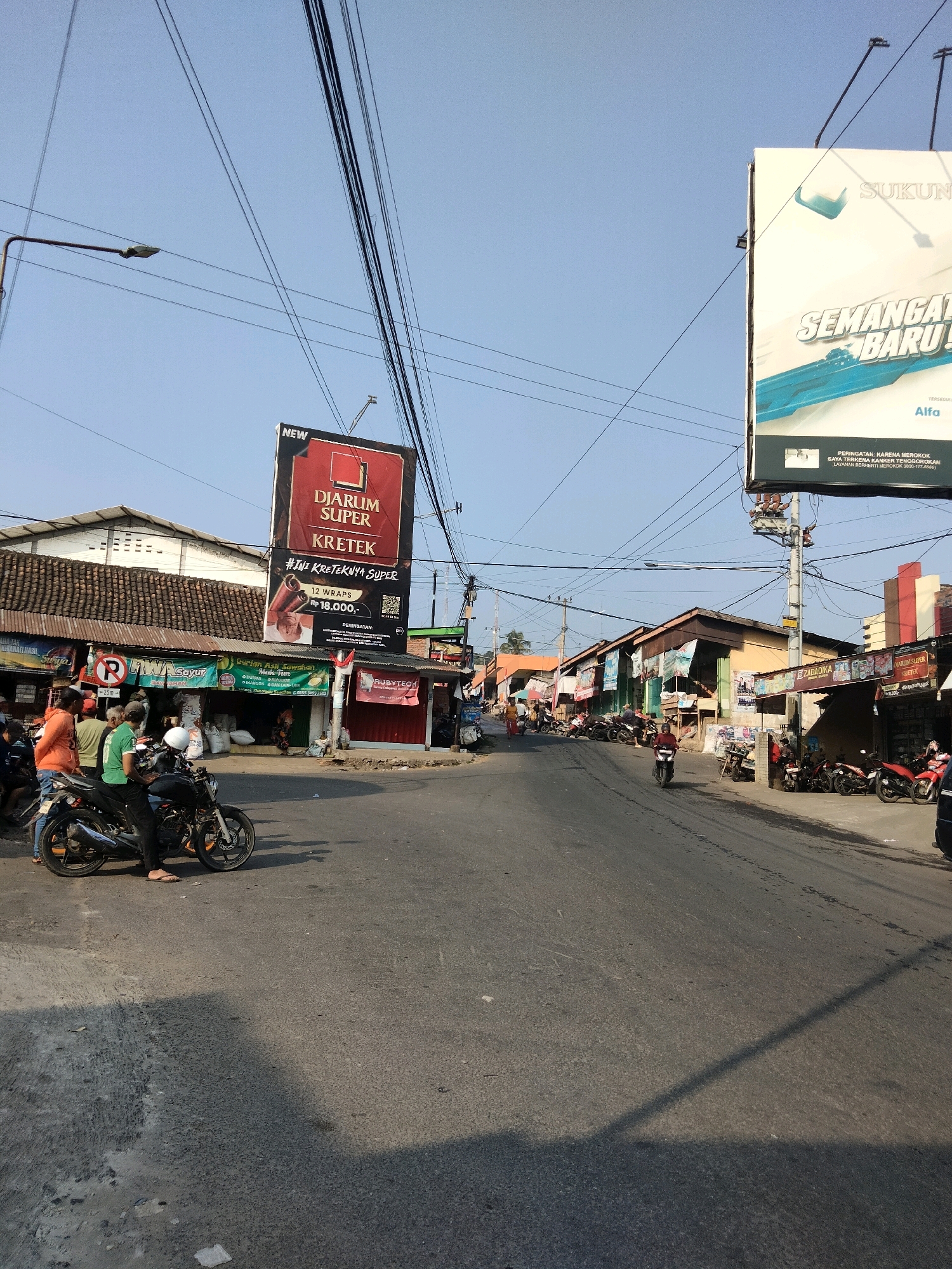 The image shows a street scene in Sawahan, Indonesia, likely a market or commercial area. The sky is clear and blue. Overhead, numerous electrical wires crisscross the frame. On the left side of the street, a tall billboard with a red and black advertisement for "DIARUM SUPER KRETEK" is prominently displayed. Below the billboard, a row of businesses with corrugated metal roofs line the street. Some of these businesses appear to be shops selling various goods.
In the foreground, on the left side of the road, several people are gathered. One individual is standing next to a black motorcycle, and another is on the motorcycle. Other pedestrians are also visible. The street itself is paved and shows signs of wear and tear.
Further down the street, more shops and buildings are visible, many with awnings or tarps providing shade. Several motorcycles are parked along the side of the road. A larger billboard, mostly white with blue text and an image of what appears to be a cigarette pack, is visible on the right side of the street. This billboard also contains a health warning in smaller text. The overall impression is a busy, everyday scene in a commercial district.