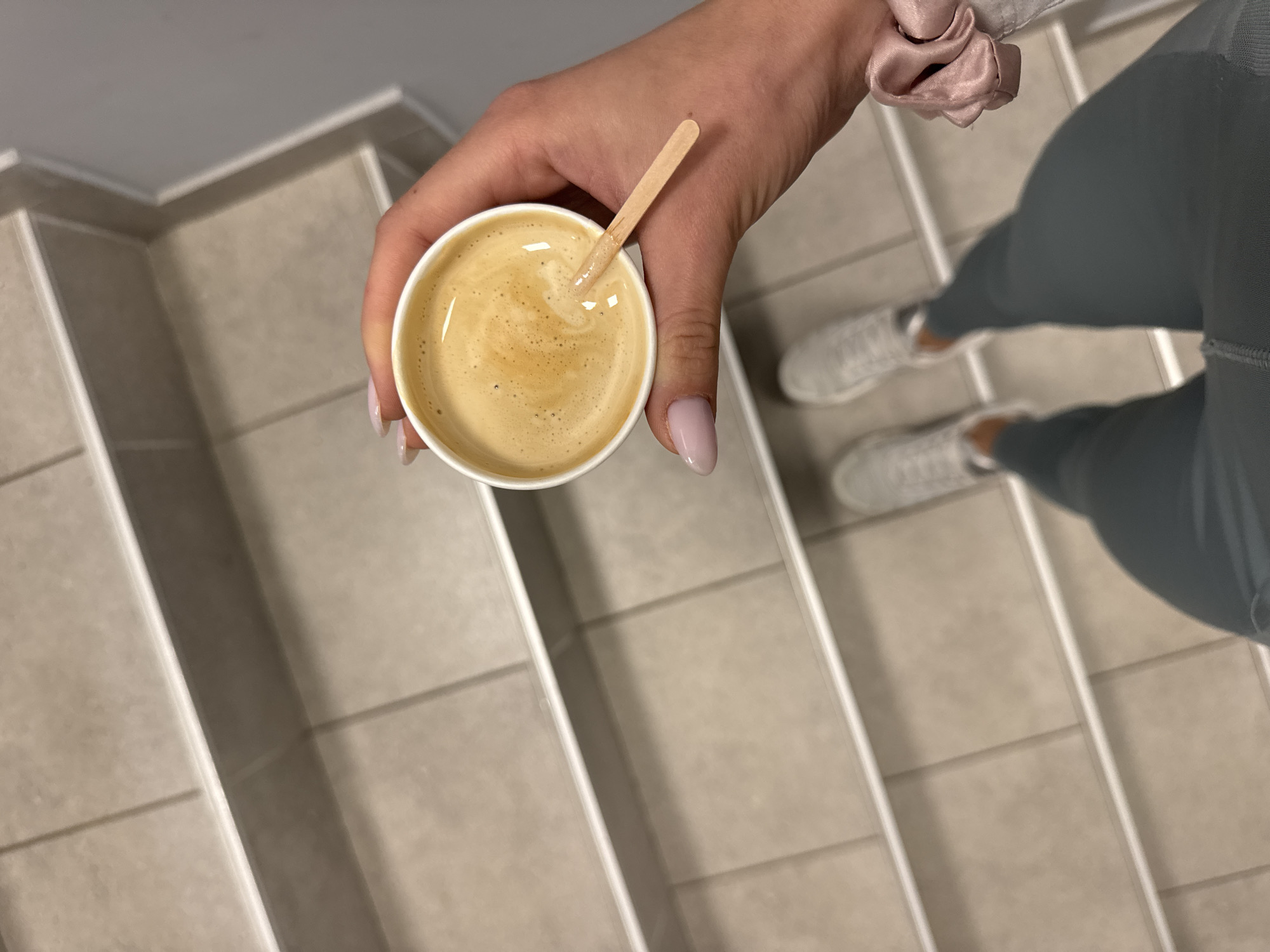 A high-angle shot depicts a hand holding a small white coffee cup with a wooden stirrer inside. The coffee has a light brown color with visible foam. The hand has pale pink nails. A pink scrunchie is visible on the wrist. The person is wearing grey leggings and white sneakers. The background consists of tiled stairs with a neutral color scheme. The location cannot be determined based on the image.