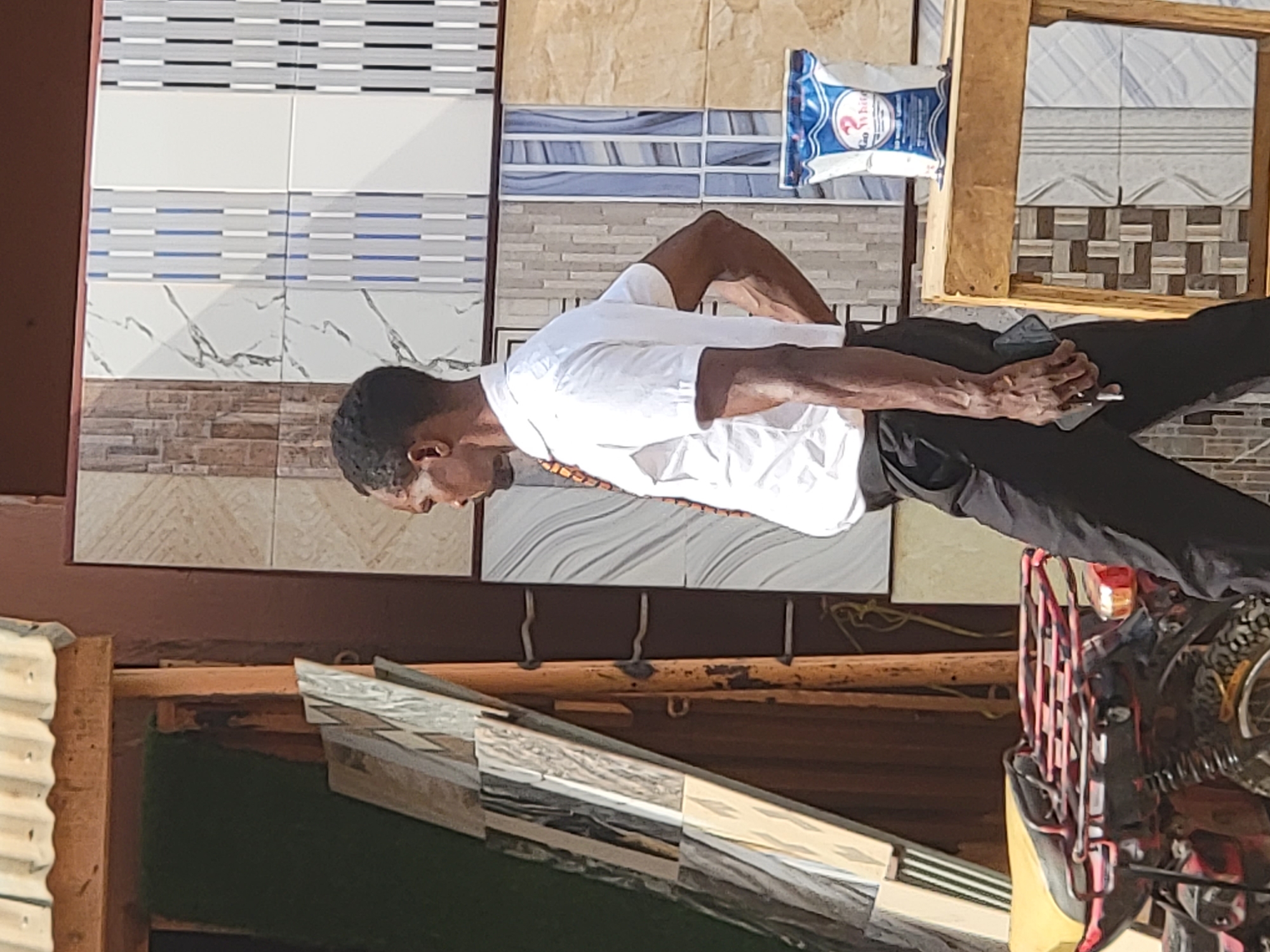 A man stands in an outdoor setting, possibly a building material shop or stall in Maiburiji, Nigeria. He is positioned in the center-left of the frame, wearing a white short-sleeved shirt and dark trousers. His hands are clasped together in front of his waist, and he is looking downwards with a pensive or focused expression. He appears to be a Black man with short, dark hair.
Behind him, a wide variety of ceramic or porcelain tiles are displayed on what looks like a wall or display board. The tiles feature diverse patterns, including plain white, horizontal stripes in grey and blue, marbling effects, brick-like textures, and geometric designs. Some tiles are arranged in larger panels, showcasing different styles.
In the upper right portion of the image, resting on a wooden frame, is a white and blue bag, possibly containing cement, grout, or tile adhesive. The word "White" is visible on the bag, along with a number "2" and potentially other unreadable text. To the right of the man and in the lower foreground, parts of a motorcycle or scooter are visible, including a red metal rack and a tire, suggesting it might be parked nearby.
The lighting indicates it is daytime, likely sunny, given the bright illumination. The scene appears to be an active commercial area where building supplies, specifically tiles, are sold. In the far left foreground, corrugated iron sheeting is partially visible, typical of market stalls or temporary structures. The wooden frames holding some of the tiles and the general setup suggest a practical, perhaps informal, retail environment.