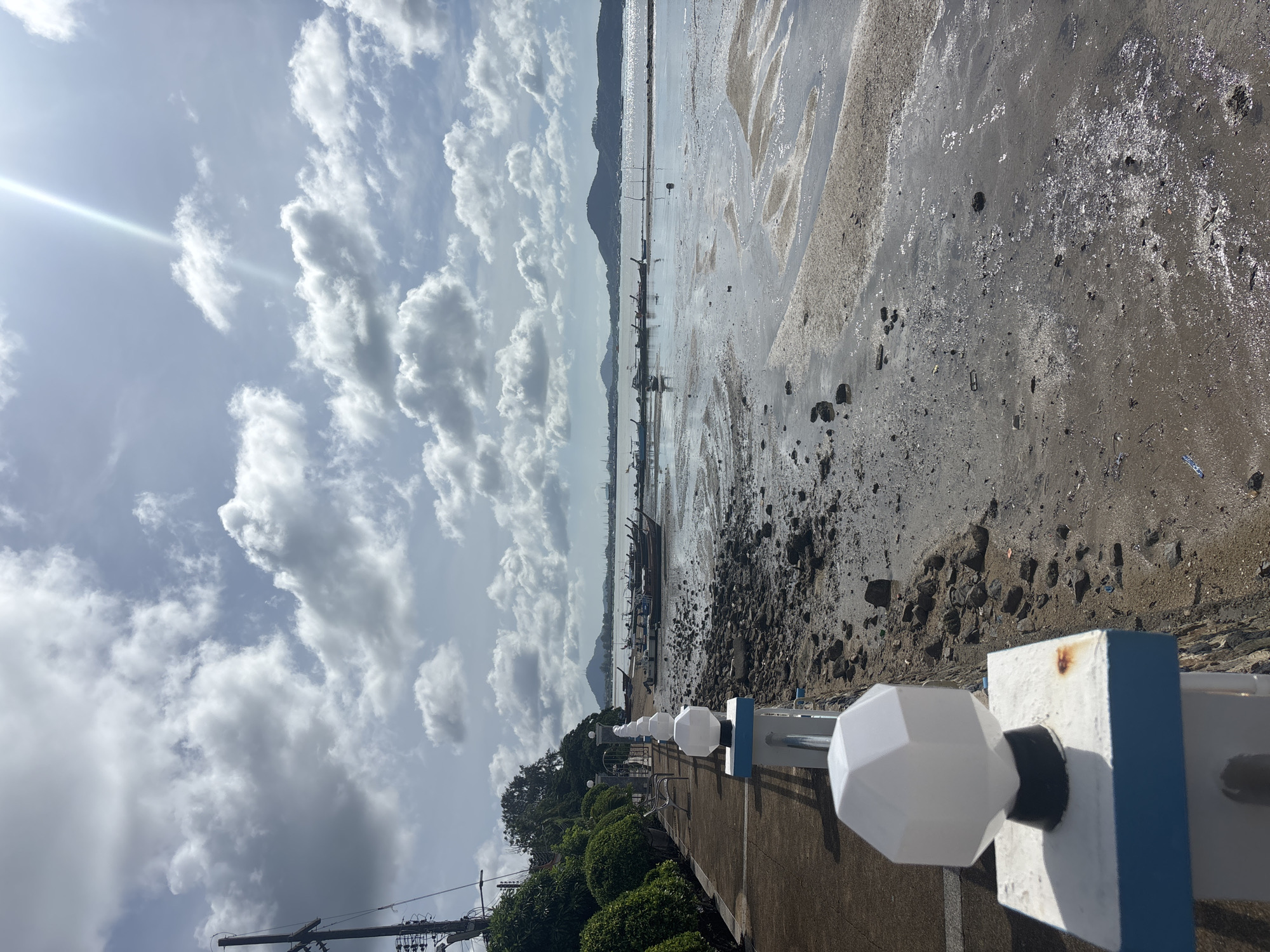 This image depicts a seaside promenade in Rawai, Thailand, under a partly cloudy sky with visible sun glare. The foreground features a concrete walkway bordered by a white railing topped with decorative light fixtures. To the right of the walkway is a muddy, rocky shoreline exposed by low tide, with scattered debris. Several long-tail boats are moored in the shallow water in the middle ground. Beyond the boats, the sea stretches to a distant, tree-covered island under a hazy sky. Lush green bushes and a metal gate are visible along the left side of the promenade, with utility poles and wires further in the background.