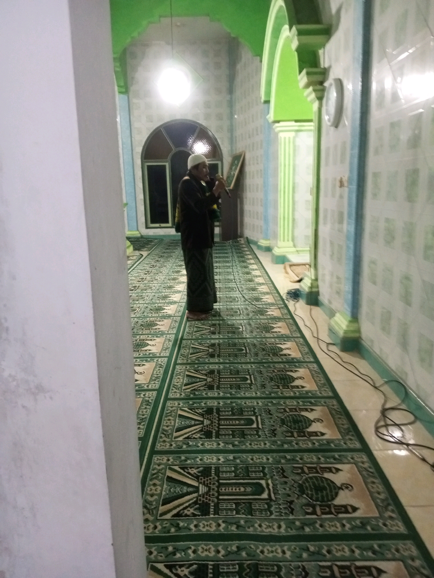 In the city of Andongsili, Indonesia, a man wearing a white skullcap and dark clothing stands in a mosque. He is holding a microphone and appears to be speaking or singing into it. The mosque has a long, patterned carpet with green and tan designs, and the walls are tiled with green and white patterns. The ceiling is painted green, and there are arched doorways leading to other parts of the building. A clock is visible on the wall. The scene is dimly lit, with a bright light source illuminating the space from above.