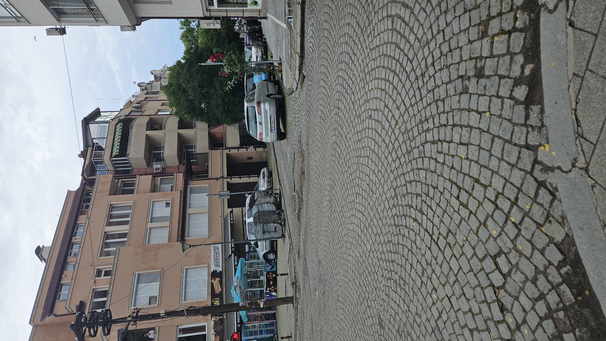 The image depicts a street scene in Plovdiv, Bulgaria. The street is paved with cobblestones in a curved pattern. A row of buildings with peach-colored facades, windows, and balconies lines the left side of the street. Cars are parked along the street. A business with "BECKS" signage is visible. A traffic light is at the bottom left. The sky is partly cloudy.