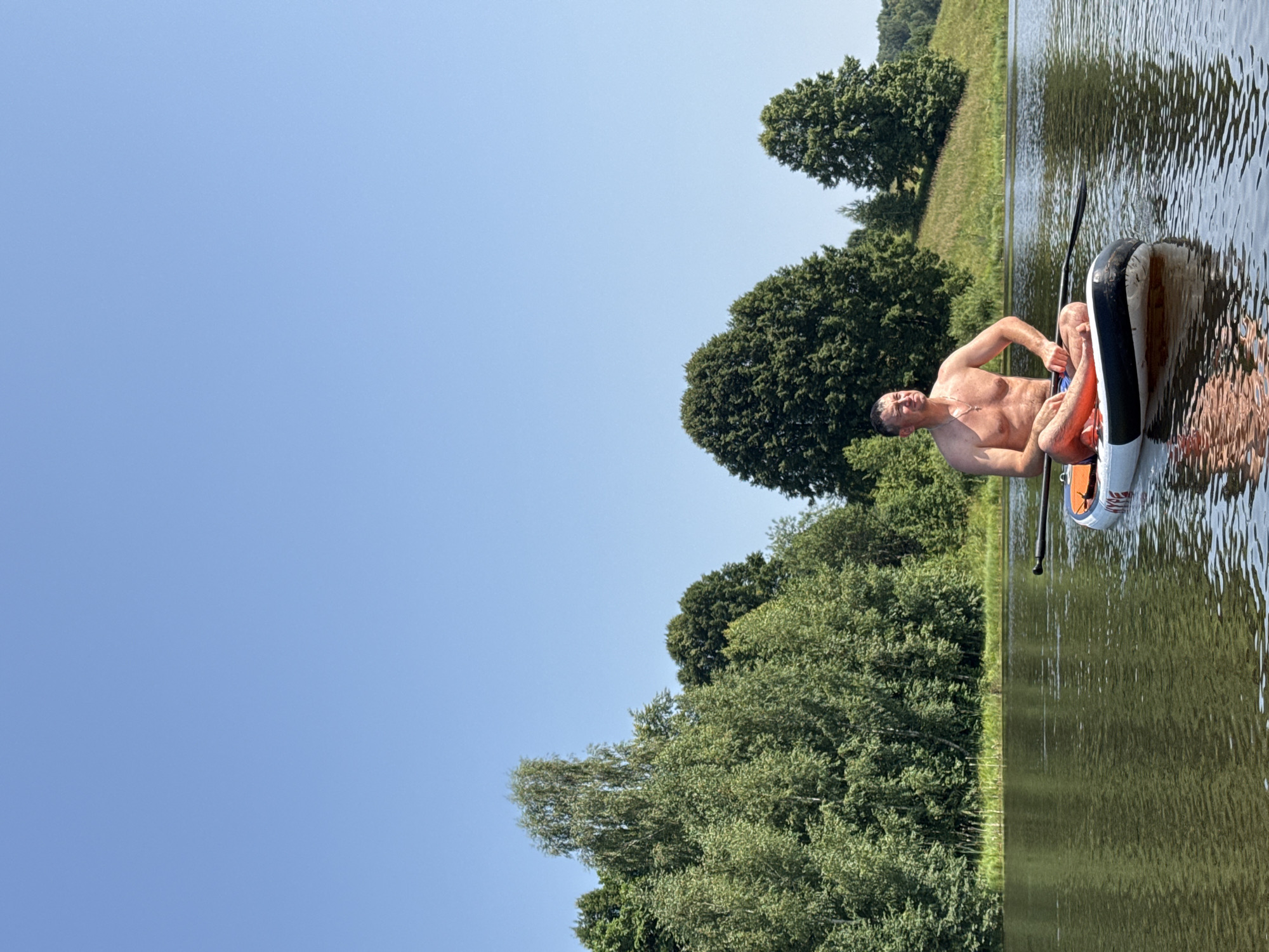 A man is paddleboarding on a calm lake on a clear day. He is shirtless and wearing blue swim trunks, and he is sitting cross-legged on the paddleboard. The paddleboard is white with orange accents and has a paddle resting across it. In the background, there is a lush green treeline with a grassy hill on the right side. The sky is a bright, cloudless blue. The location is Zasechnyi, Russia.