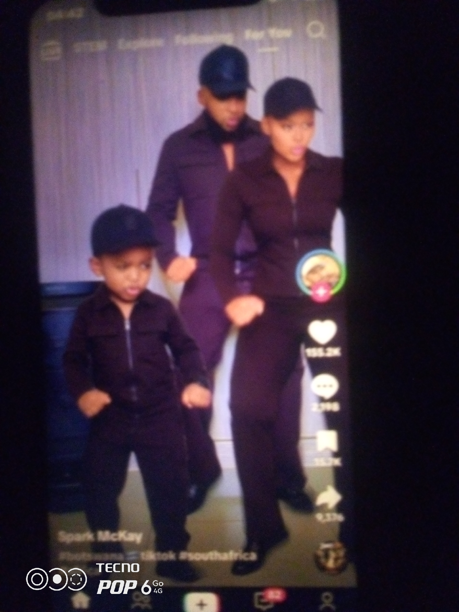 The image shows a TikTok video with three individuals. On the left, a young child wearing a black cap and a black zip-up shirt is standing with hands on hips. In the center, a man wearing a dark blue cap and a dark purple zip-up shirt stands with his head bowed. To the right, a woman in a dark blue cap and a dark purple zip-up shirt is looking forward. The individuals appear to be dancing or posing. The background is a plain, light-colored wall. On the right side of the screen, social media engagement icons are visible, including a heart with "155.2K" below it, a speech bubble with "2,198" below it, and a share icon with "9.3K" below it. At the bottom of the screen, text indicates "Spark McKay," "Whichwana," "TikTok," and "#southafrica." A "TECNO POP 6 Go 4G" watermark is in the lower left corner. The image is a low-resolution, likely captured from a mobile phone screen. The location is not specified as Howayi, Nigeria.