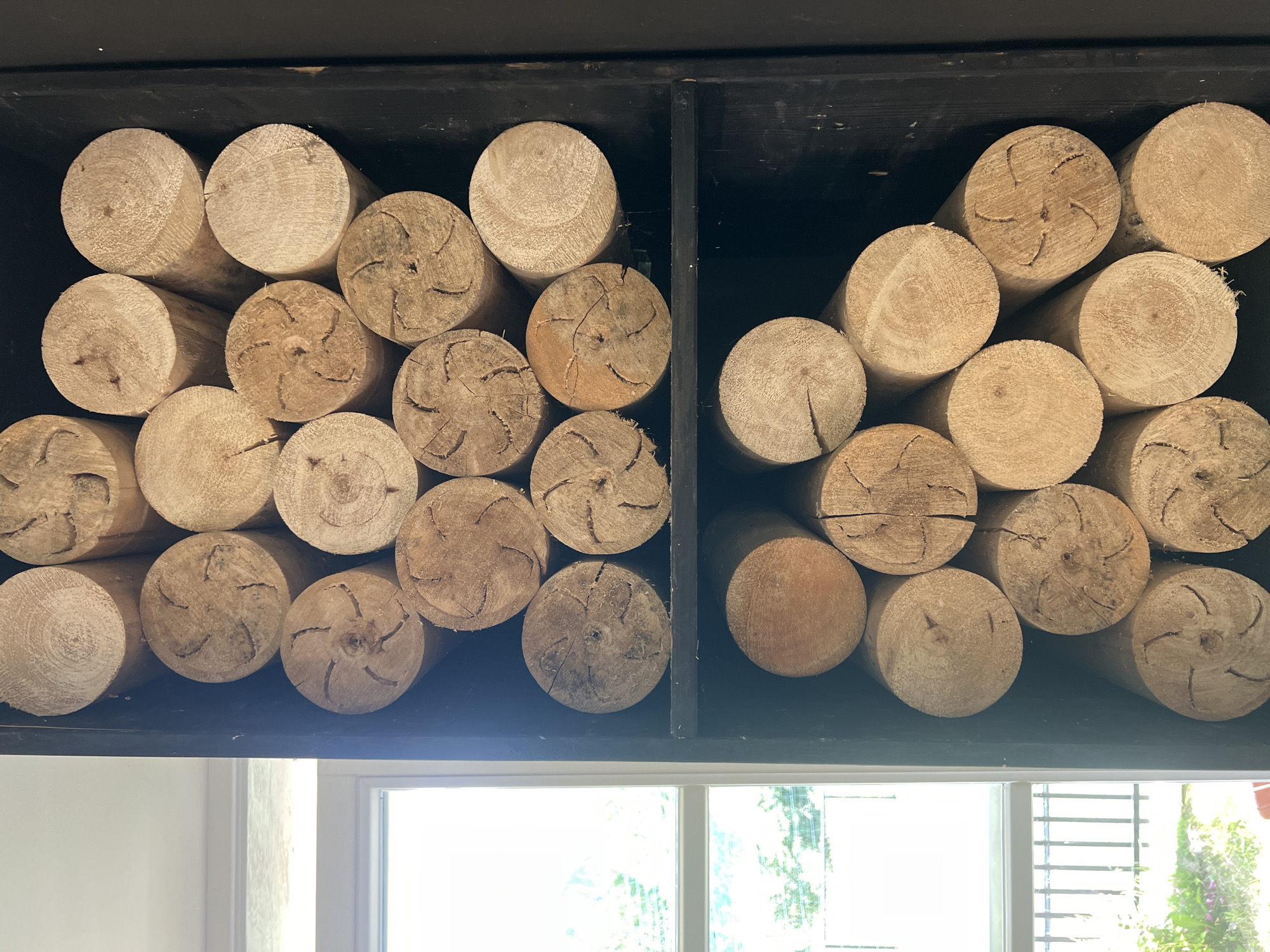 A close-up, vertical shot captures a dark wooden shelving unit filled with stacked, round wooden logs. The logs are arranged in a dense, organized manner, filling the frame from top to bottom. The texture of the wood is prominent, showcasing grain patterns and the raw, cut ends of the logs. Some logs display visible cracks and unique patterns from the wood's natural growth. To the left of the shelving unit, a portion of a window is visible, offering a blurred view of greenery and a fence outside, suggesting an outdoor environment adjacent to the interior setting. The lighting appears natural, casting subtle shadows and highlighting the wood's texture.