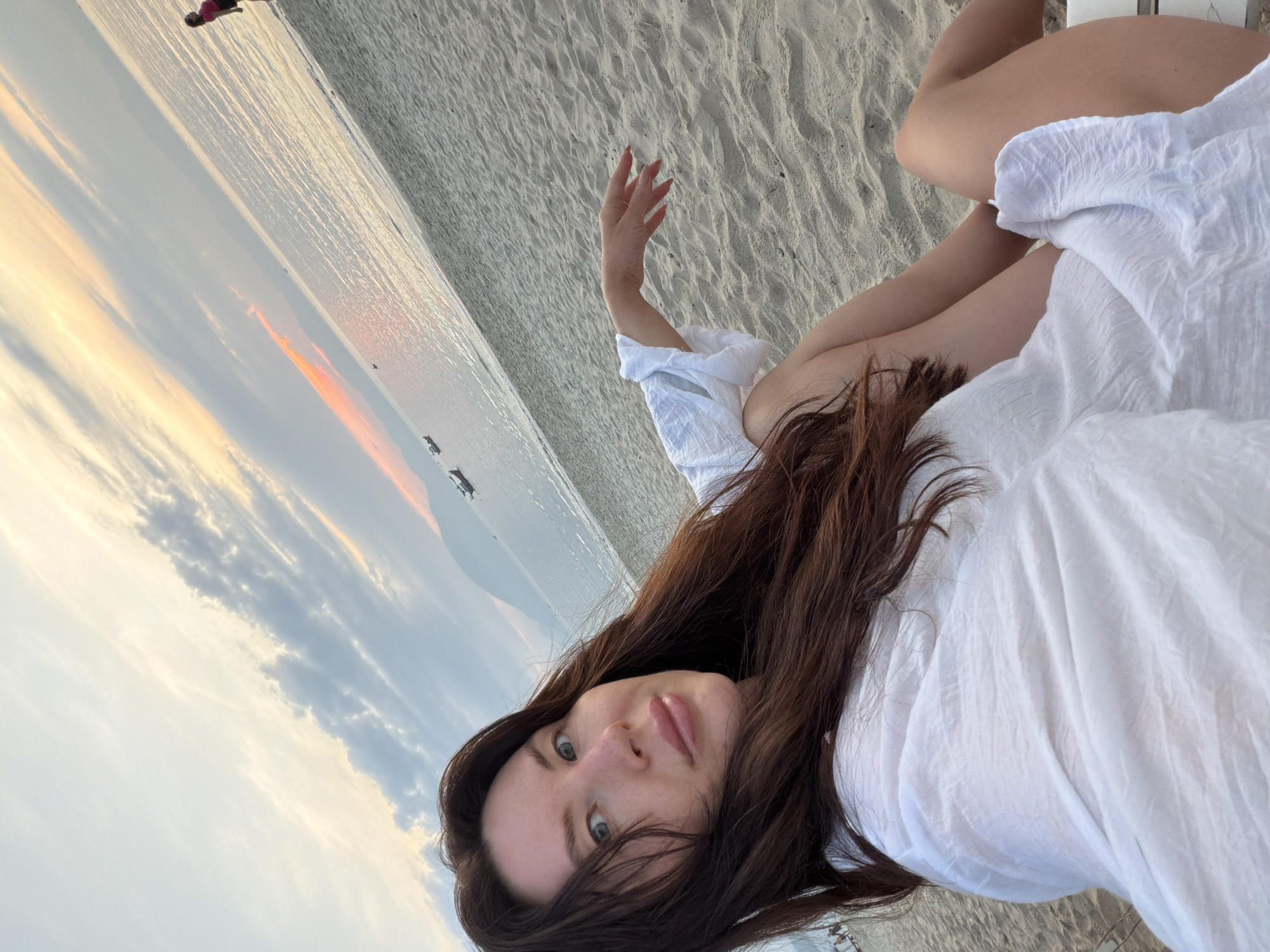 A woman with long, dark hair sits on a beach chair on a sandy beach, facing the camera. She is wearing a white, sheer top. Her left arm is resting on her lap, and her right arm is extended with her hand open. In the background, the ocean meets the sky at sunset, with soft orange and pink hues in the clouds. There are a few small boats on the water and a faint outline of mountains in the distance. To the far right of the image, a person in a pink shirt and black shorts is standing on the beach. The scene is located in Ninh Hoa Ward, Dong Nai Province, Vietnam.
