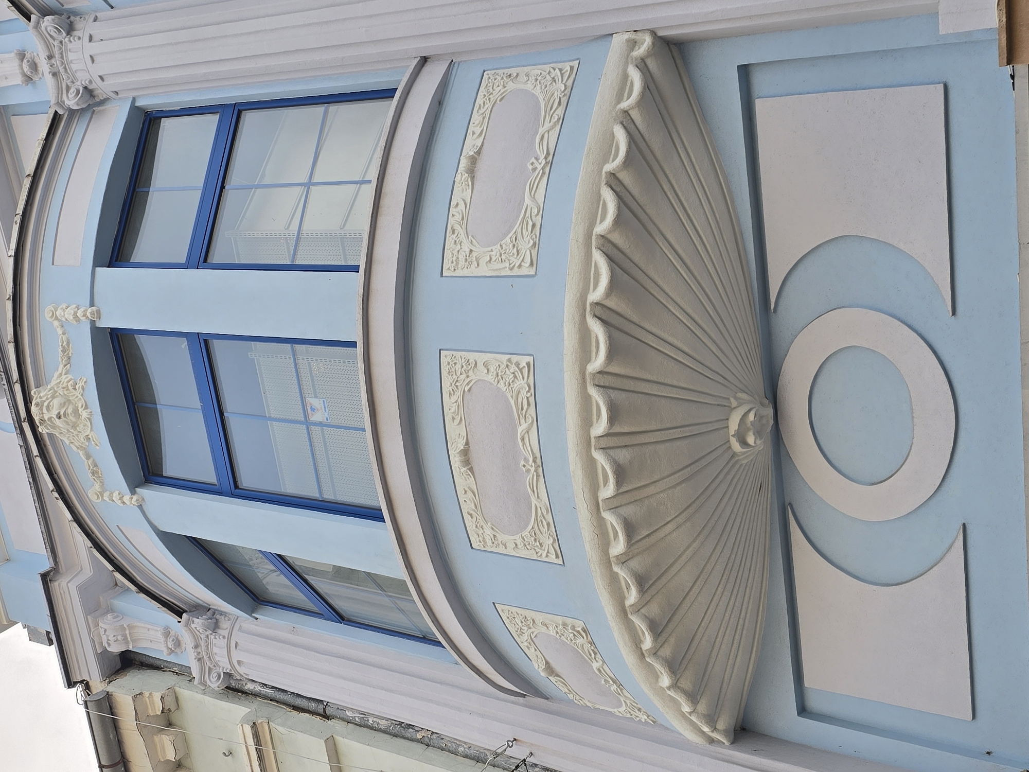 A low-angle, medium-shot, color photograph displays a building’s exterior wall in Unknown, Unknown. The wall features a curved section with windows, decorative plasterwork, and a pale blue facade.
Three blue-framed windows are visible. Decorative plaster frames in a pale tone are visible between the windows and on the facade’s side. A sculptural element resembling an open fan or shell-like design is prominently displayed, composed of fine ribs radiating from a central point.
To the right of the fan-like structure is a design composed of a rectangle above a circular form that is placed on top of the rectangle, all rendered in white plaster. The background wall is painted a light blue hue. Additional architectural details are visible at the top and bottom of the frame. The overall aesthetic evokes an impression of classical or historical design.