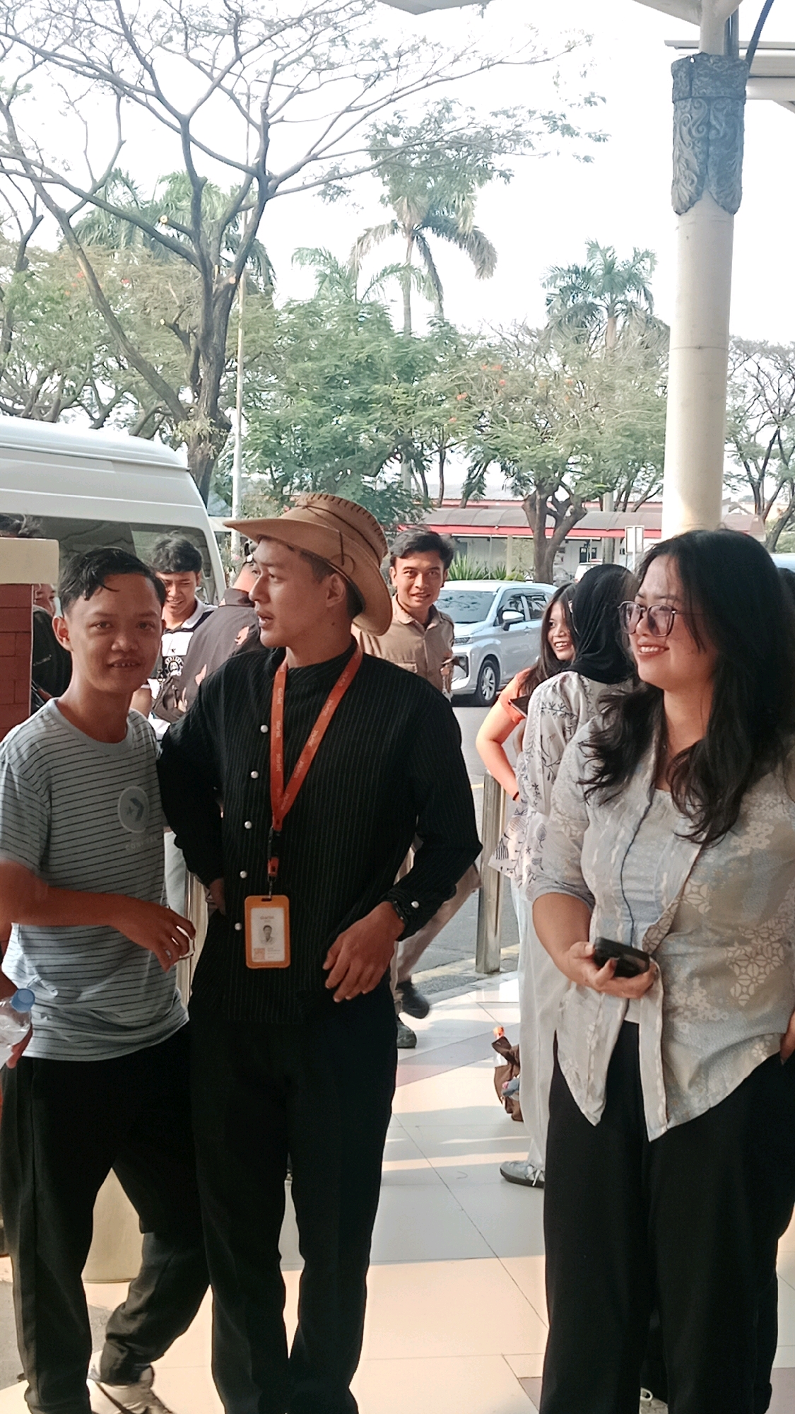 An outdoor medium shot shows a group of people standing near a white van and a building. The person in the foreground on the left is a man wearing a striped t-shirt and black pants. To his right, a man in the center is wearing a black, pinstriped long-sleeved shirt, black pants, and a wide-brimmed brown hat. He has an orange lanyard with an ID card around his neck. To the right of the man in the hat, a woman in a light-colored patterned shirt and dark pants is looking to her left and holding a phone in her right hand. In the background, other individuals are visible, including a man in a light brown collared shirt and a woman wearing a dark headscarf. Trees and palm trees are visible behind the group. The scene appears to be in an urban setting, possibly near an entrance or parking area. The presence of palm trees suggests a tropical or subtropical climate. The overall atmosphere appears casual and possibly a gathering or event.
