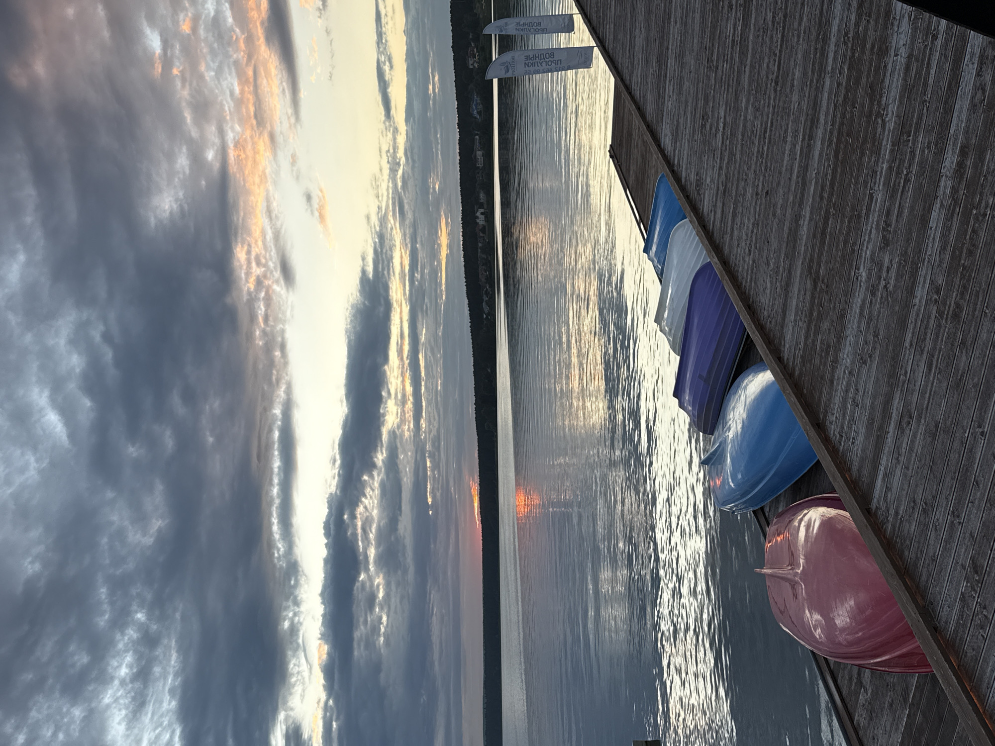 The image depicts a serene waterscape at dusk on a lake, with a wooden pier in the foreground. Several small boats, in shades of red, blue, and purple, are neatly lined up and overturned on the pier. Two white flags with Cyrillic text stand upright on the pier, suggesting a recreational or event location. The water is calm, reflecting the muted colors of the sky and the distant tree line. The sky itself is filled with dramatic clouds, with a sliver of orange and pink from the setting sun breaking through the darker gray tones. The overall impression is one of tranquility and natural beauty, potentially at a resort or park area in Votkinsk, Russia, given the Cyrillic text on the flags.