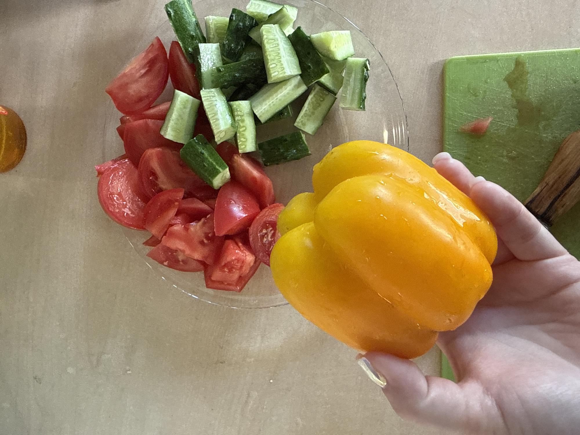 In this overhead shot, a hand holds a bright yellow bell pepper, its skin glistening with water droplets. The pepper is positioned in the foreground, partially obscuring a clear glass plate filled with chopped vegetables. Sliced red tomatoes and cut green cucumbers are arranged on the plate, appearing to be prepared for a salad.
To the right of the plate, a vibrant green cutting board is visible, also wet, suggesting recent food preparation. A wooden utensil rests on the cutting board. The background is a textured surface, likely a table, in a neutral, light brown tone.
The lighting is bright and natural, casting soft shadows, and there are no discernible time of day or weather cues. The setting appears to be a kitchen or dining area. The focus is on the fresh ingredients and the process of preparing food. No text is visible in the image. The location context provided indicates this scene is in Nizhne-Chufichevo, Russia.