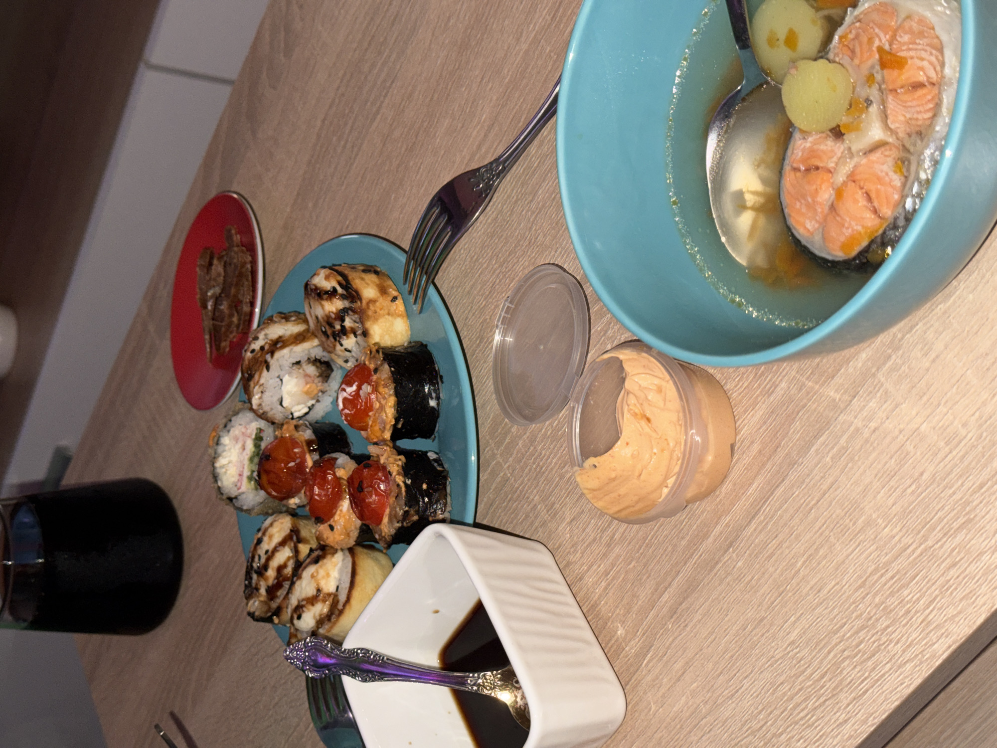 The scene depicts an indoor dining setting on a light-colored wood grain tabletop, in the city of Unknown, Unknown. A blue circular plate displays multiple sushi-style rolls; some are baked with white toppings and black sesame seeds, while others are topped with whole red cherry tomatoes. A smaller red circular plate holds several dark brown, thinly sliced meat pieces. A light blue bowl contains clear broth, a pink salmon steak, and several small, light-colored spherical items, with a metal spoon partially submerged. A white rectangular ramekin contains a dark brown liquid and a decorative metal spoon. A small, clear plastic container with a partially open lid holds an orange-colored creamy substance. A dark glass bottle or carafe is positioned in the upper left background. A metal fork with a decorative handle rests on the tabletop. The background consists of light-colored furniture or cabinetry.