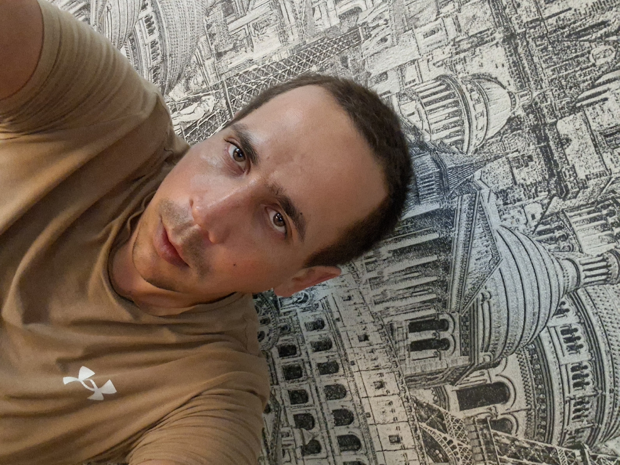 This is a selfie taken by a man in his 30s with short brown hair and brown eyes, looking directly at the camera. He is wearing a light brown t-shirt with a white Under Armour logo on the left side of his chest. The background is a wall with a black and white pattern featuring architectural elements like domes, columns, and the Eiffel Tower, suggesting a Parisian theme. The lighting is dim, and the man's face appears slightly shiny, indicating sweat. There are no other people or objects clearly visible in the frame. The man's expression is neutral.