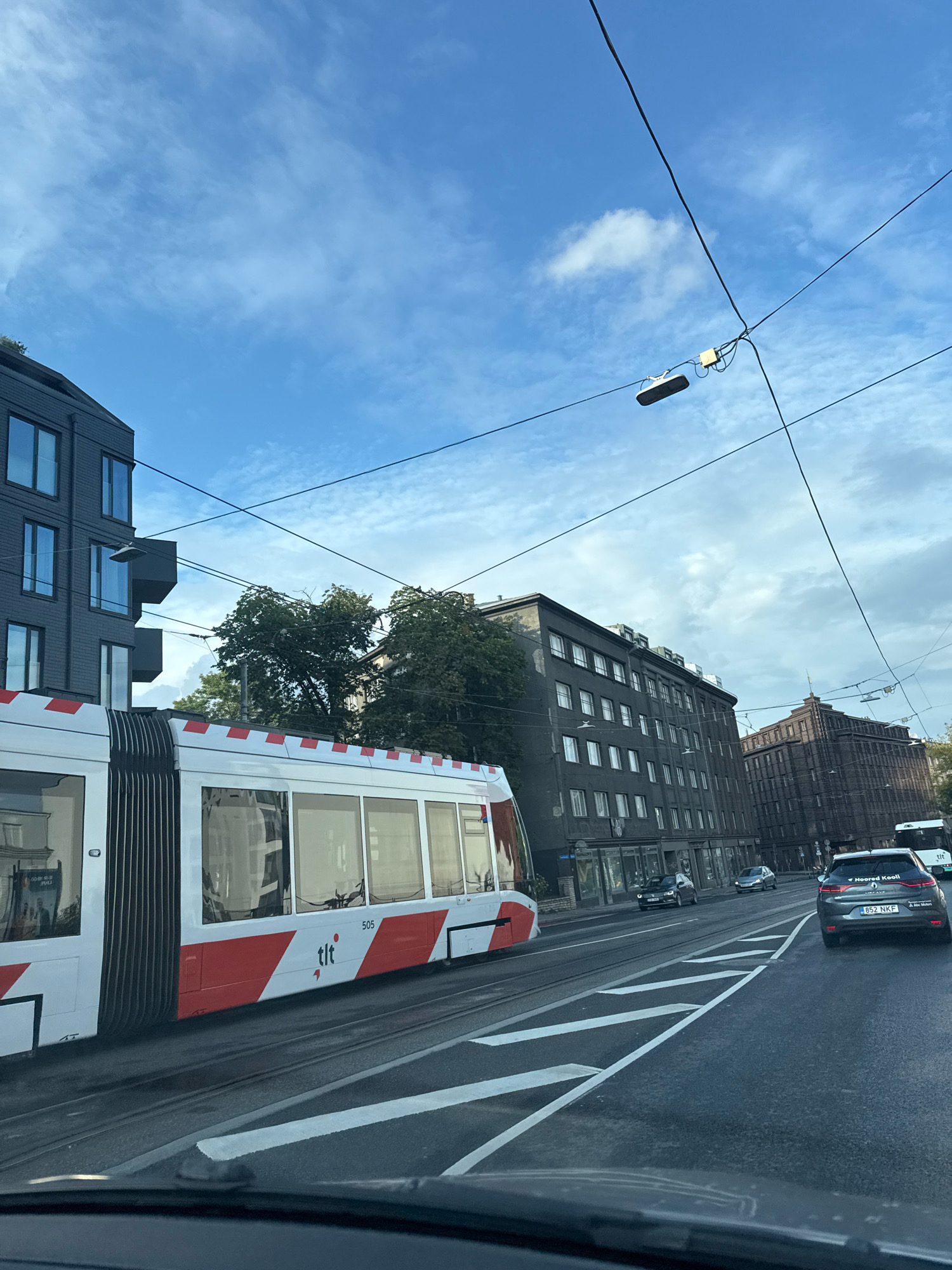 A street-level, eye-level, exterior, vertical, medium shot captures a public transport tram, a modern dark gray building with reflective windows, and a dark brick building on a cloudy day in Tallinn, Estonia. The tram, predominantly white with red accents and the "tlt" logo, is on tracks in the foreground, positioned on the left side of the frame. The tram has the number 505 visible on its side. Behind the tram, a four-story modern building with dark gray siding and large windows stands to the left. Further to the right, a multi-story dark brick building with numerous windows is visible. The street features lane markings and traffic, including a dark gray car with license plate "852 NKF" in the right lane, and another vehicle partially visible in the distance. Overhead, power lines crisscross the sky, with a light fixture attached to one of them. Trees with green foliage are present between the buildings. The sky is a mix of blue with scattered white clouds. The overall scene depicts urban transportation infrastructure and architecture.