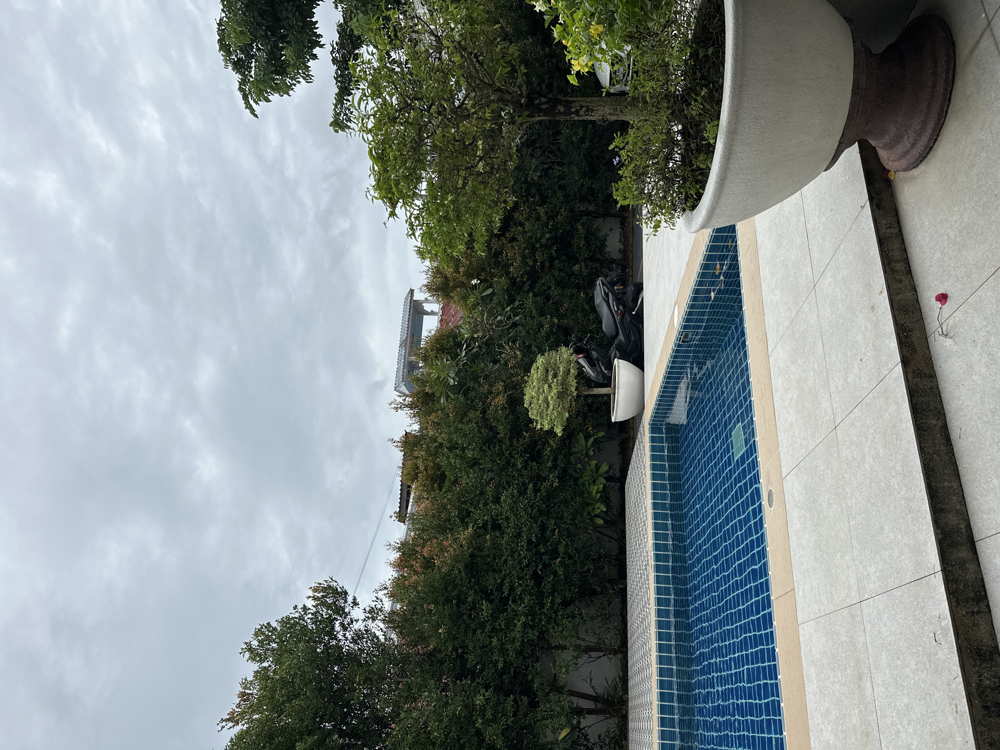 The image depicts an outdoor swimming pool area on an overcast day. The pool itself is rectangular with blue tiled walls and a light-colored coping. The surrounding patio is paved with large, light gray tiles. To the left of the pool is a dense hedge of dark green bushes, with some reddish new growth visible at the top. Further back, behind the hedge, are glimpses of buildings with red roofs. In the middle ground, to the right of the pool, a black motorcycle is parked next to a white pot containing a small, rounded tree. To the far right, a large, decorative planter holds a tree with green foliage, and some yellow flowers are visible in the background. The sky is filled with gray, textured clouds.
