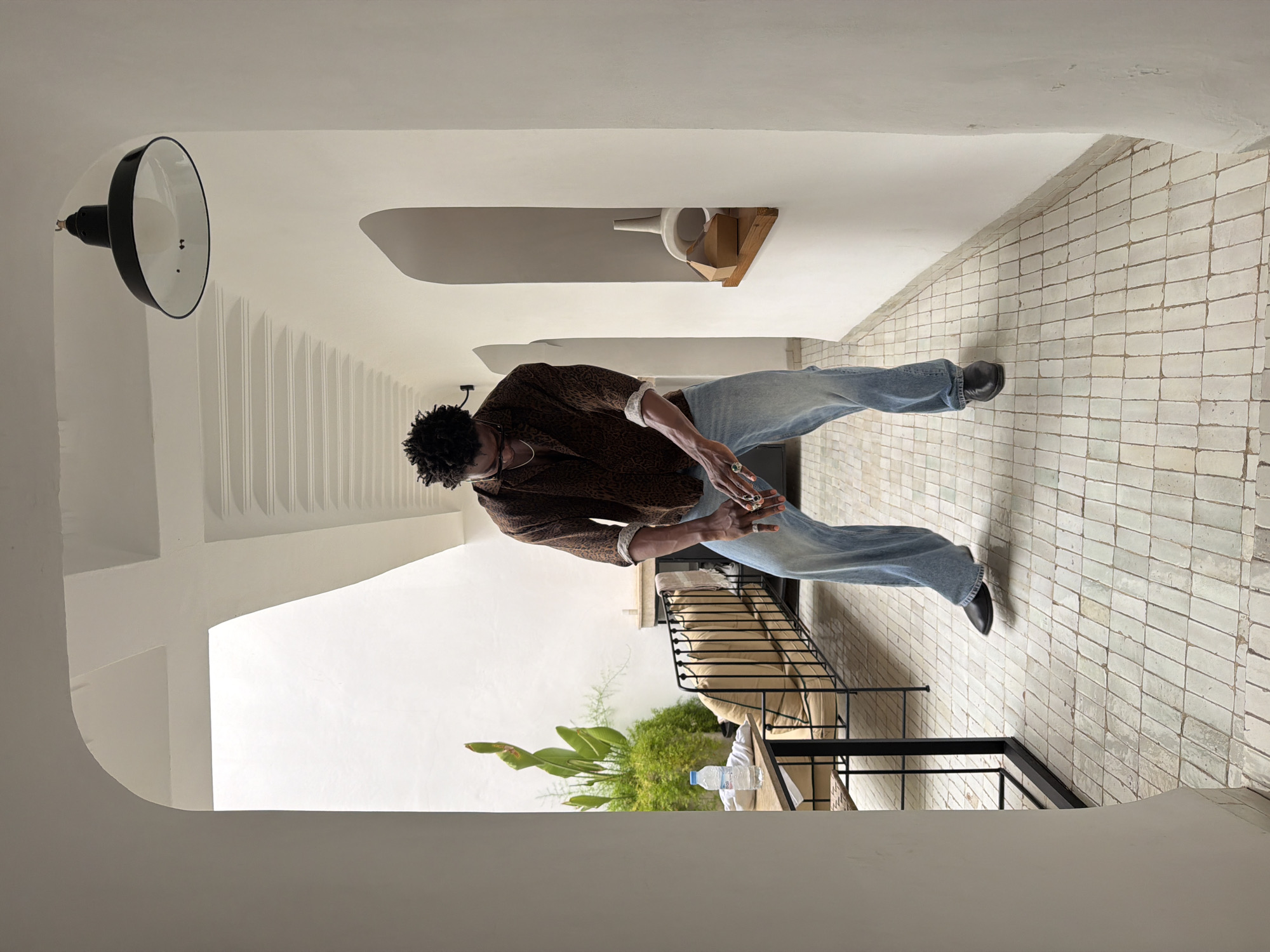 The image depicts an outdoor corridor with white stucco walls and a tiled floor. A Black male, wearing a patterned brown button-down shirt, light-wash baggy jeans, and black loafers, is the central subject. He is bending forward with his hands close together, appearing to be examining something in his palms. To the left of the corridor, an outdoor seating area is visible, featuring a lounge couch with beige cushions and a metal frame, a small table with a water bottle and white cloth, and lush green plants. The architecture suggests a Riad in Marrakesh, Morocco, characterized by its white walls, arched openings, and decorative ceiling elements. A black industrial-style pendant lamp hangs from the ceiling in the upper right quadrant of the image. The recessed niche on the right wall contains two ceramic vases and a small brown box on a wooden shelf. The lighting is bright and diffused, indicating daytime.
