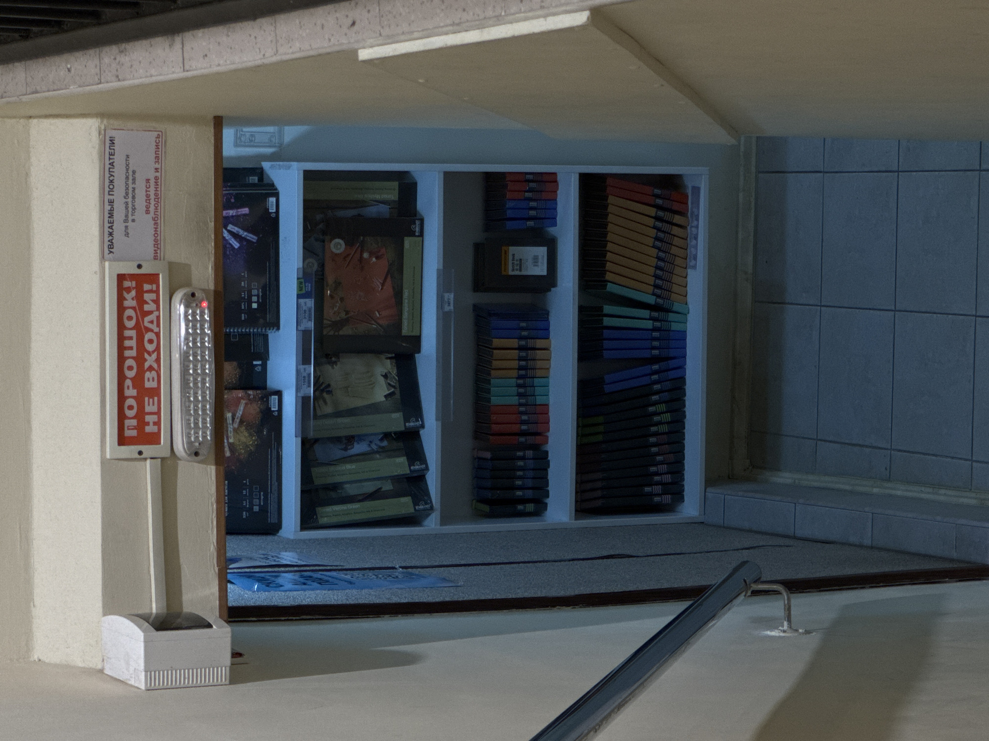 The image shows an interior view of what appears to be a storage area or a small shop. The scene is dimly lit, suggesting it might be nighttime or a poorly lit indoor space. The overall impression is somewhat cluttered, with shelves filled with numerous items, predominantly video game cases or similar media.
In the foreground, to the left, there is a white pillar with two red and white signs. One sign reads "ПОРОШОК! НЕ ВХОДИ!" (POWDER! DO NOT ENTER!). Below this, another sign in Russian states, "УВАЖАЕМЫЕ ПОКУПАТЕЛИ! для Вашей безопасности в торговом зале ведется видеонаблюдение и запись" (DEAR CUSTOMERS! For your safety, video surveillance and recording are conducted in the sales area). Adjacent to the signs is a silver, elongated object with a red light at the top, possibly an emergency light or an alarm. A small white device with a ribbed front is on the floor beneath the signs.
The background reveals white shelving units filled with rows of stacked media cases, predominantly in shades of blue, red, and yellow. Some individual cases are visible, with titles like "Toned Mysbos" and "NEW" printed on them. The items are neatly arranged, indicating an organized storage system. The walls behind the shelves appear to be tiled with large, gray squares, and there is a subtle hint of a ceiling light source. A metal railing runs diagonally across the bottom right of the image, suggesting the presence of stairs or a walkway. The overall atmosphere is functional and perhaps a bit utilitarian, with no people visible. The location is identified as Moscow, Russia.
