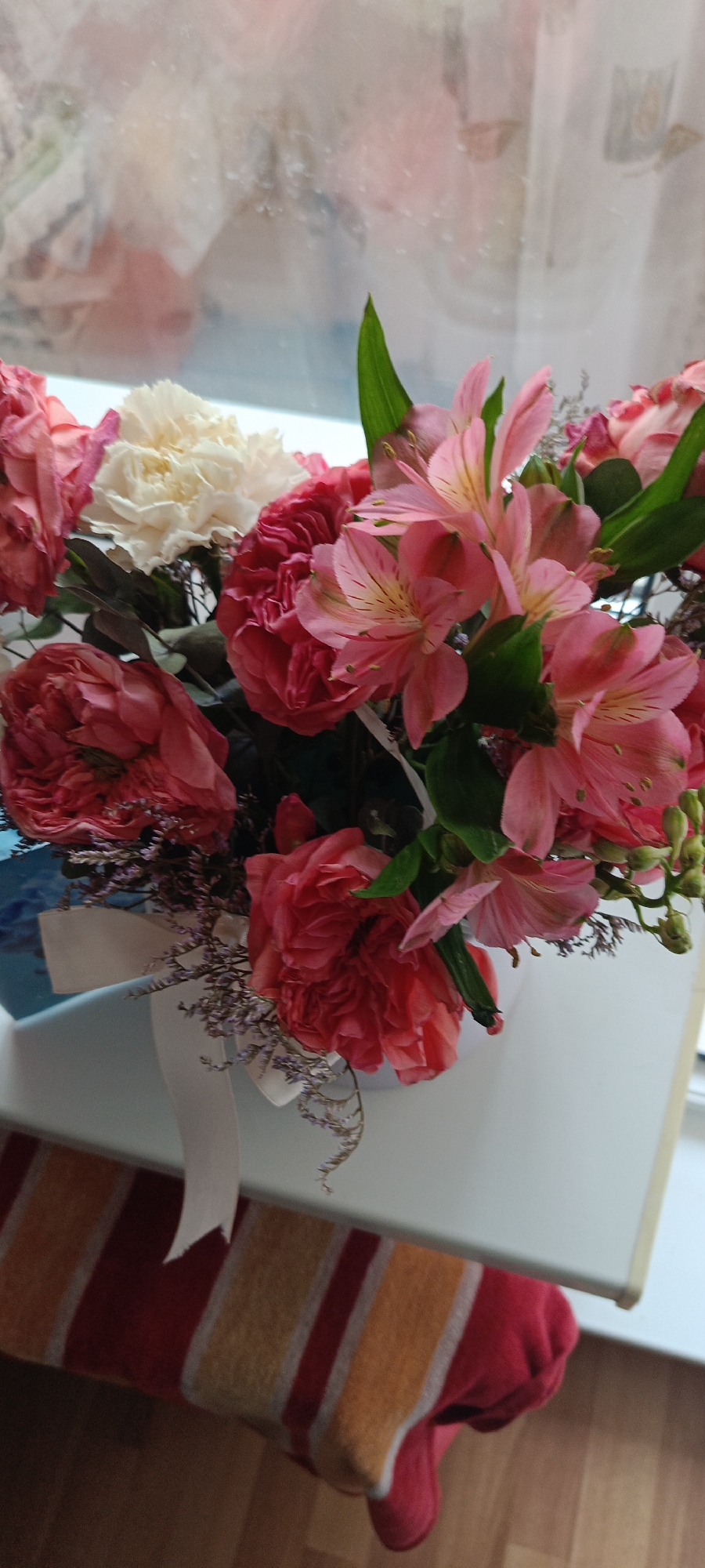 A bouquet of pink and white flowers is displayed in a white box tied with a white ribbon. The box sits on a white surface next to a window. In the background, through the window, there is a blurred reflection of what appears to be a street or outdoor scene. Below the white surface, a striped textile item, with bands of red, yellow, and white, is visible. The floor beneath appears to be wooden. The scene suggests an indoor setting, possibly a home or shop. The city of Cheboksary, Russia, is not discernible from the visual information provided in the image.