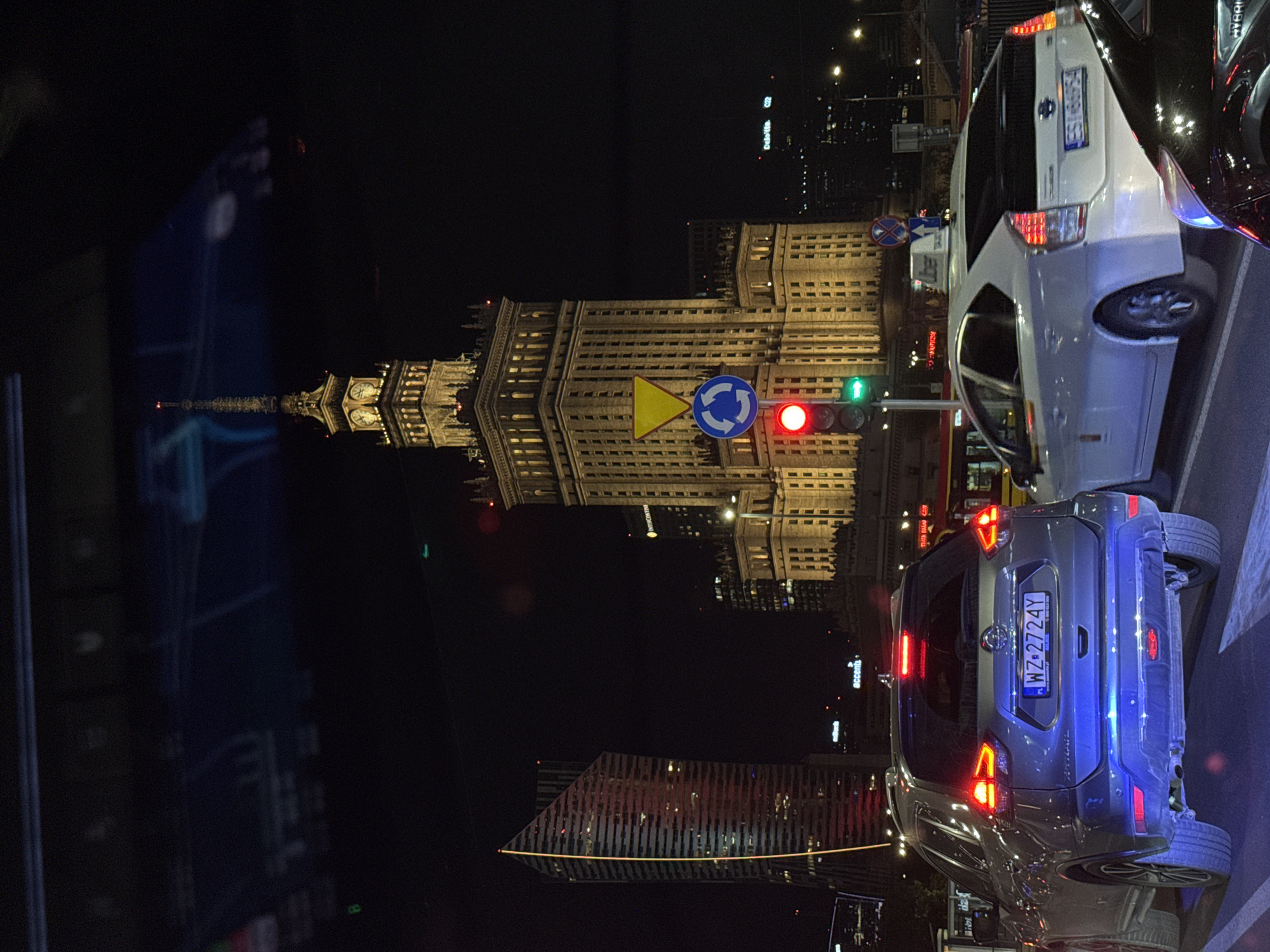 The image shows an urban scene at night. A large building with a spire, illuminated with yellow light, stands prominently in the background. Street signs, including a roundabout sign and a yield sign, are visible near a traffic light that is red. In the foreground, two cars are visible. A silver-gray SUV with a license plate reading "WZ 2724Y" has its taillights on, and a white car with a license plate reading "EST-68654" is parked nearby. The environment is dark, with other buildings and streetlights visible in the distance. The location is Warsaw, Poland, due to the visible license plates and the architecture of the large building, which is the Palace of Culture and Science.